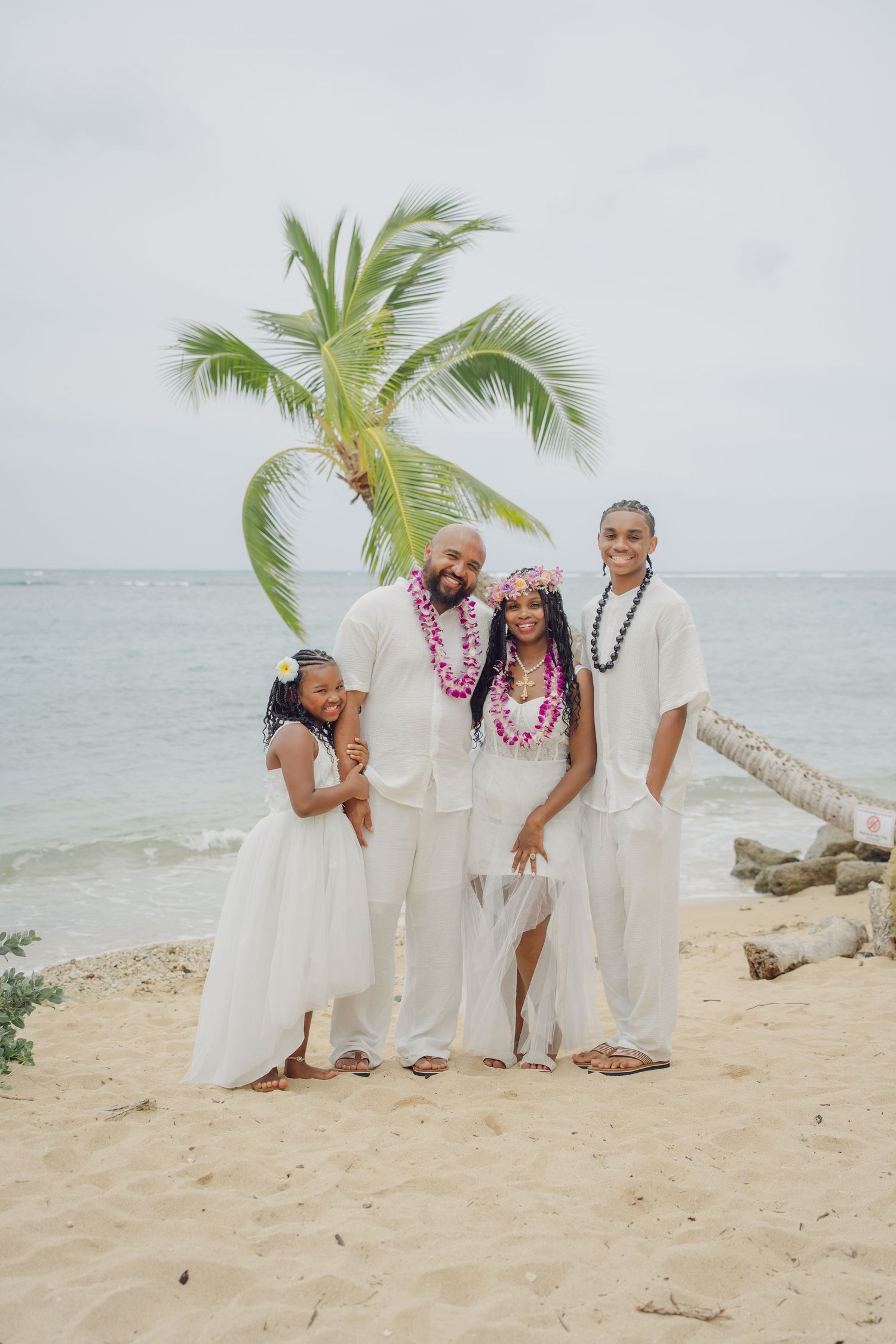 Family wearing white outfits poses together on a tropical beach with palm trees and ocean in the background.