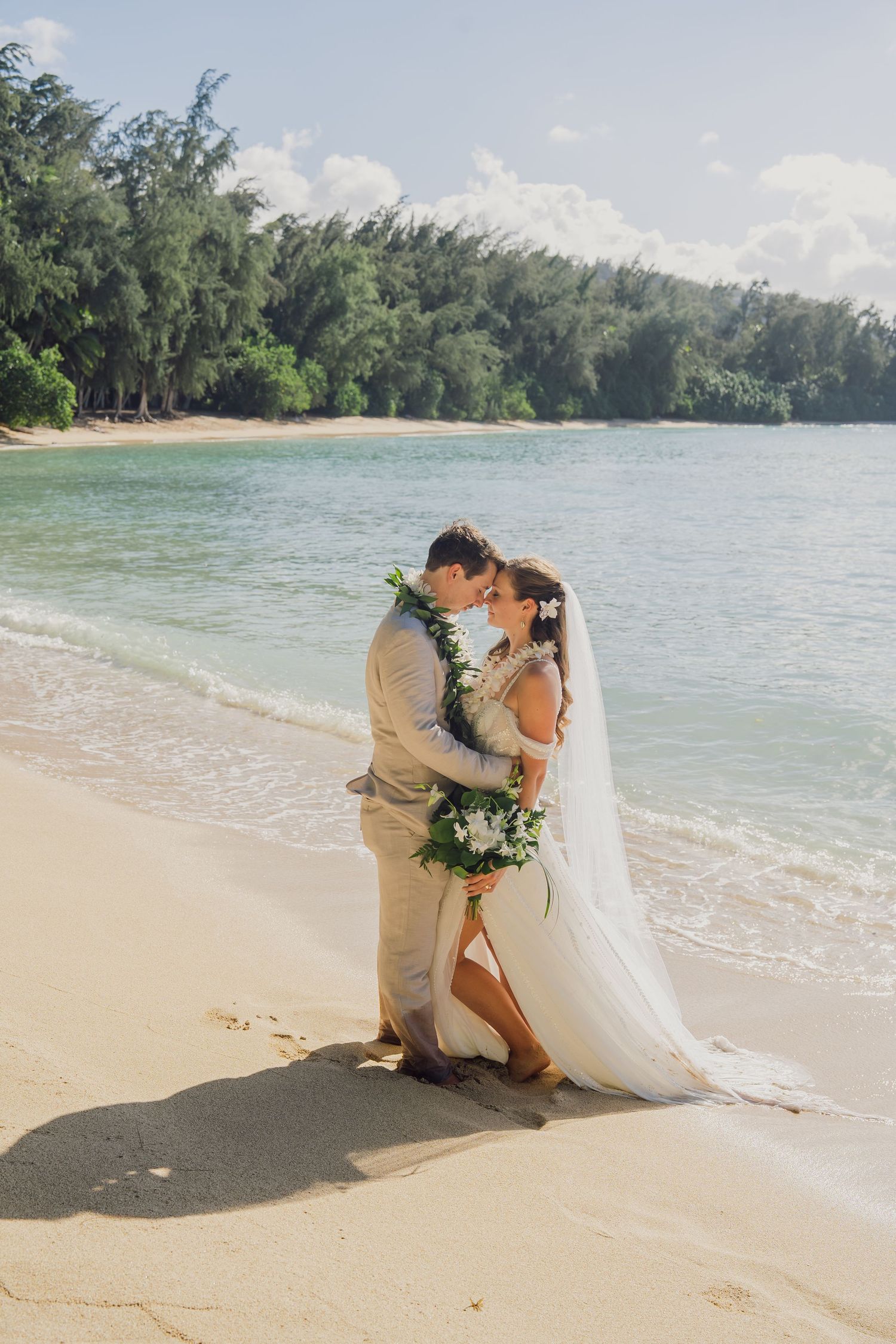 A romantic beach wedding scene unfolds with a couple embracing on a sandy shore with tropical waters and trees in the background.