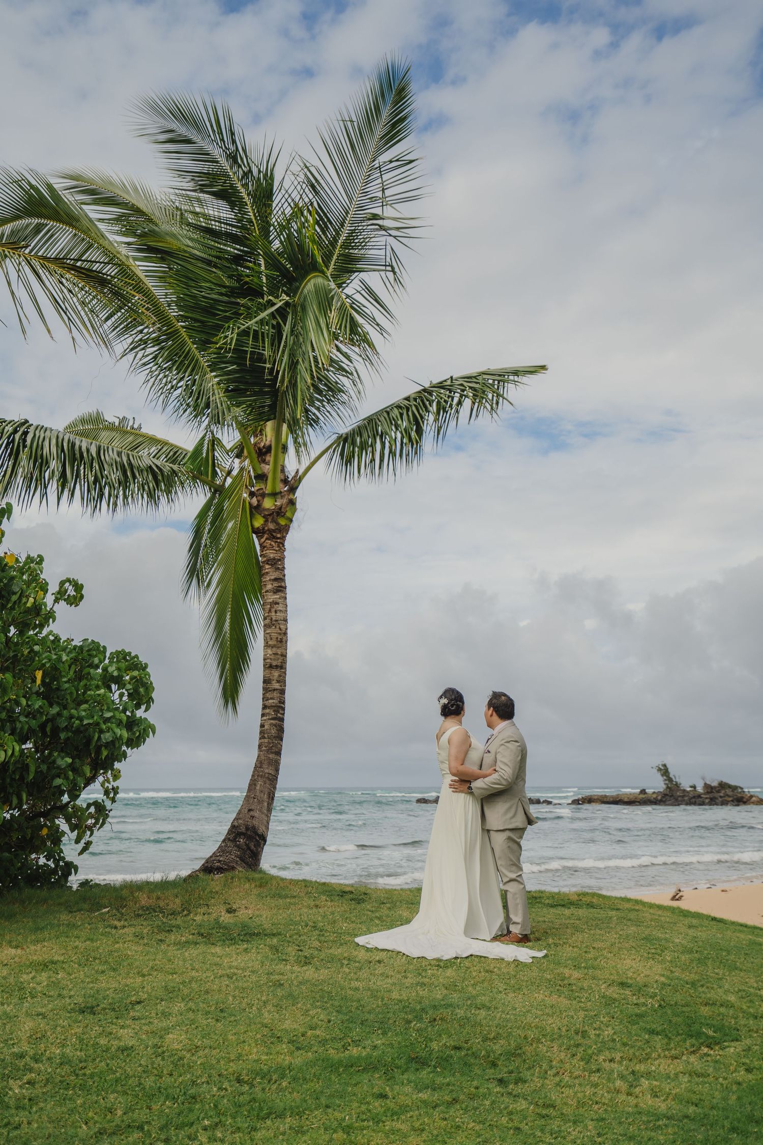 A couple stands together on a scenic beach lawn with a tall palm tree and ocean waves in the background.