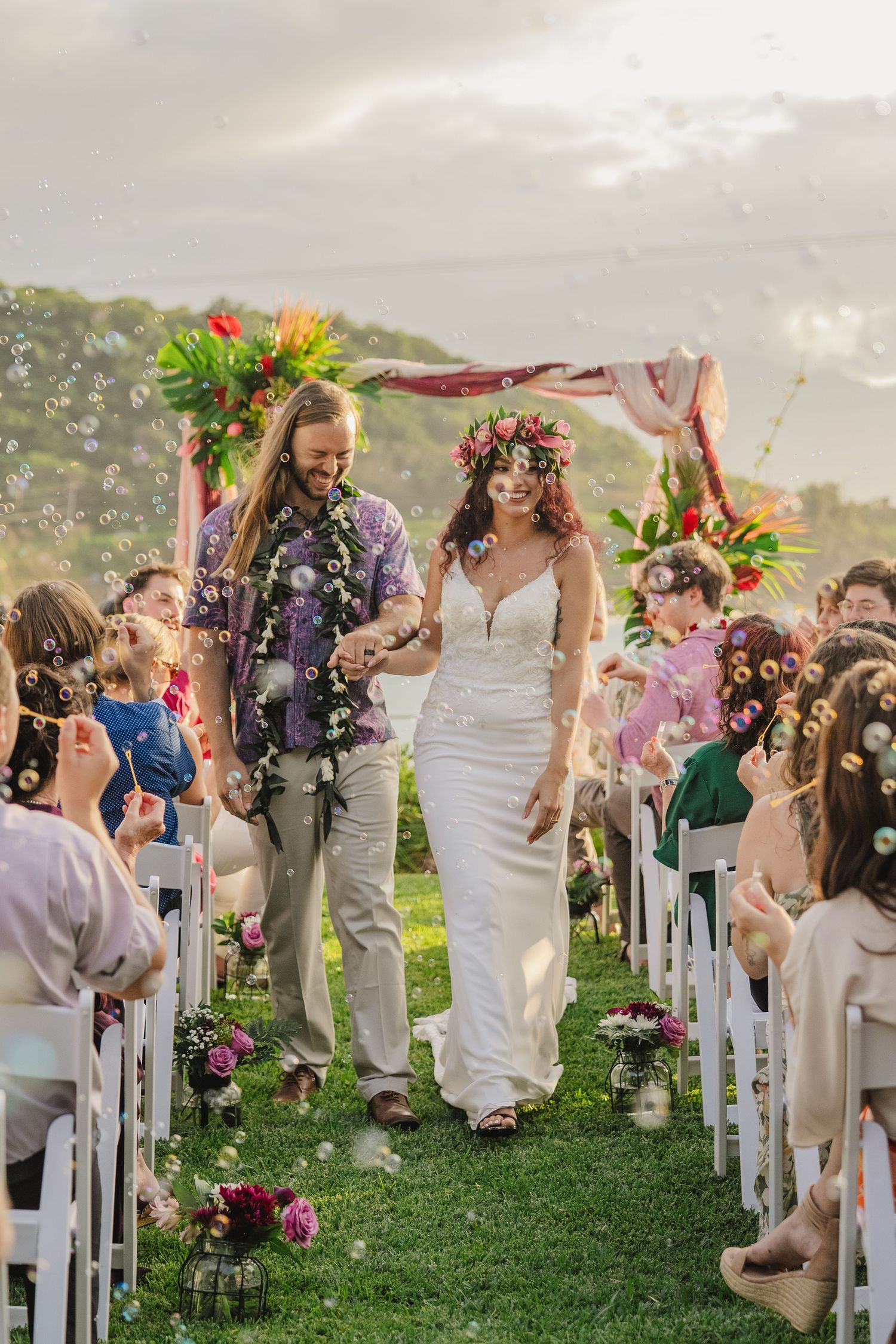 Newlyweds walk down aisle under floral arch as guests shower them with confetti at outdoor garden wedding ceremony.