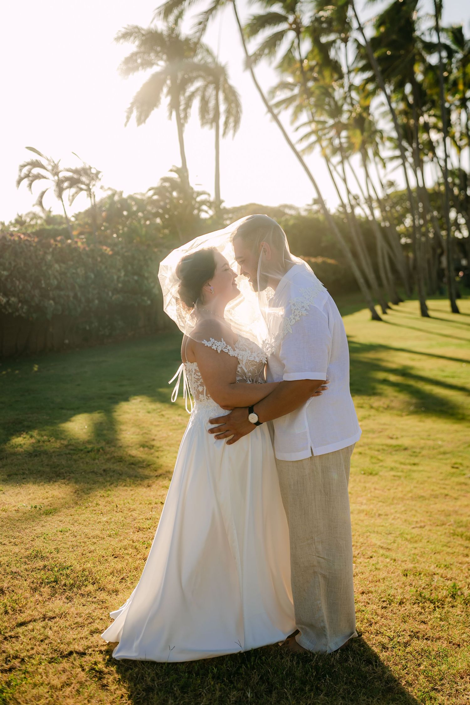 Bride and groom share a romantic kiss at sunset surrounded by tropical palm trees during their outdoor wedding ceremony.