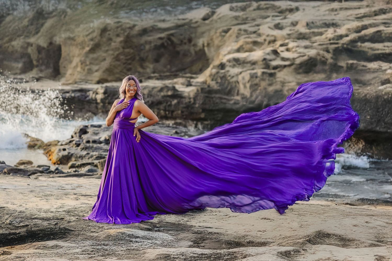 A flowing purple gown billows dramatically against rocky coastal cliffs and crashing ocean waves.