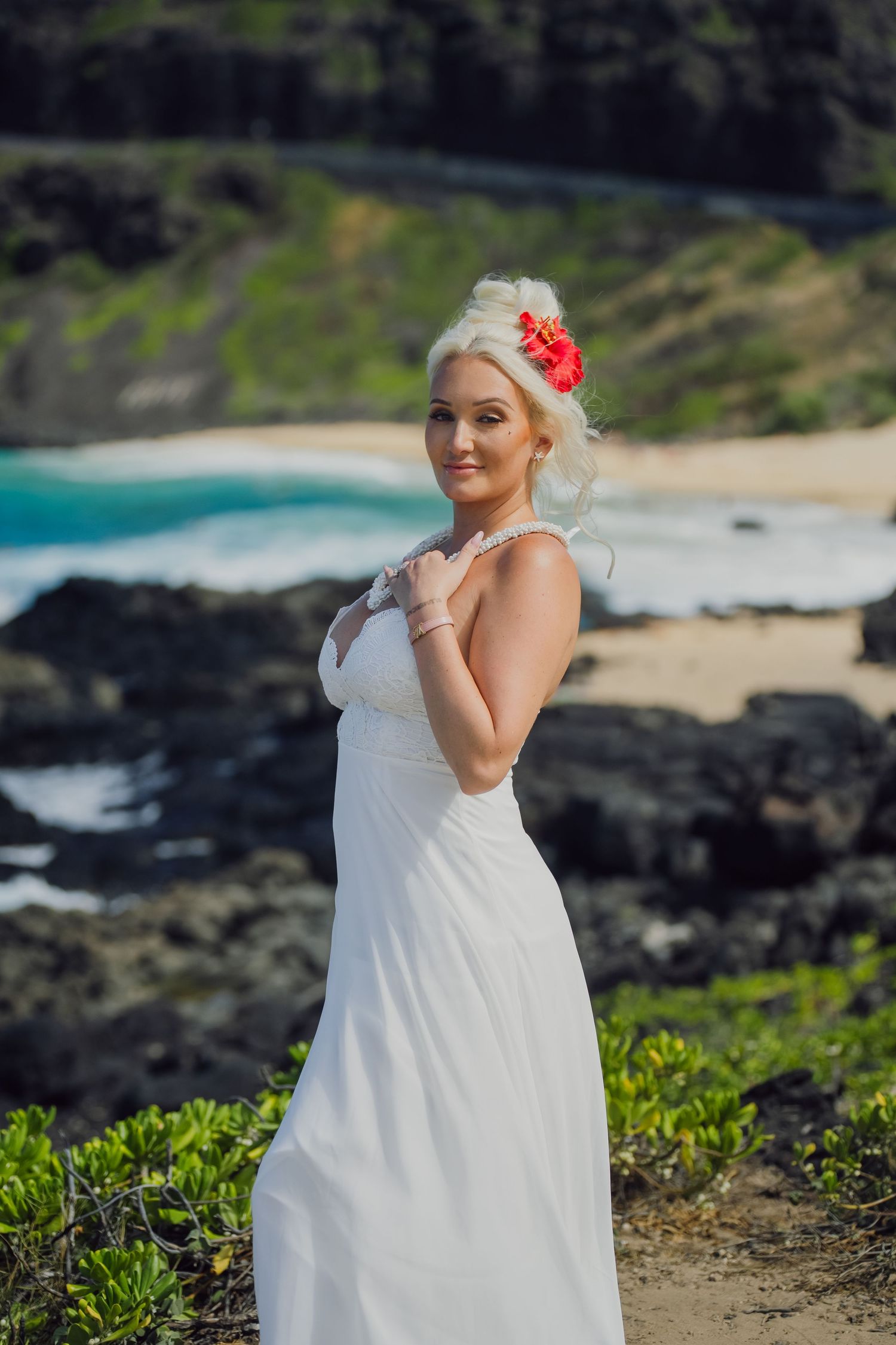 Blonde bride in white dress with red flower hair accessory posing on rocky Hawaiian beach with turquoise ocean backdrop.