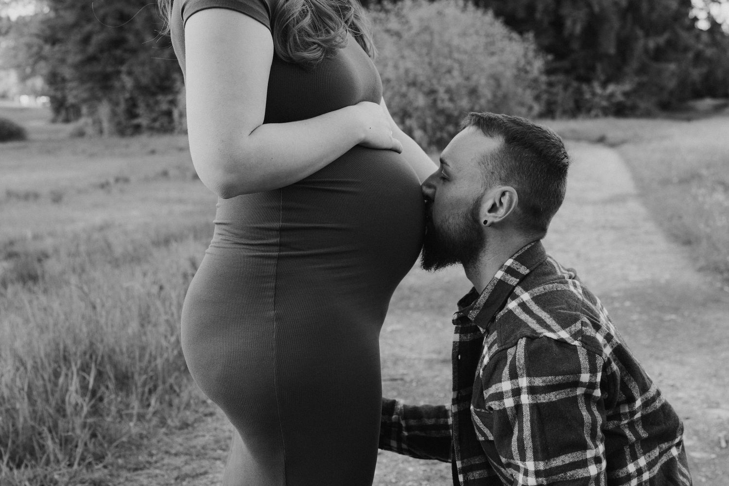 Werdender Vater kniet im Grünen und küsst den Babybauch seiner Partnerin. Emotionale Schwangerschaftsfotos im Chiemgau bei capthevibe