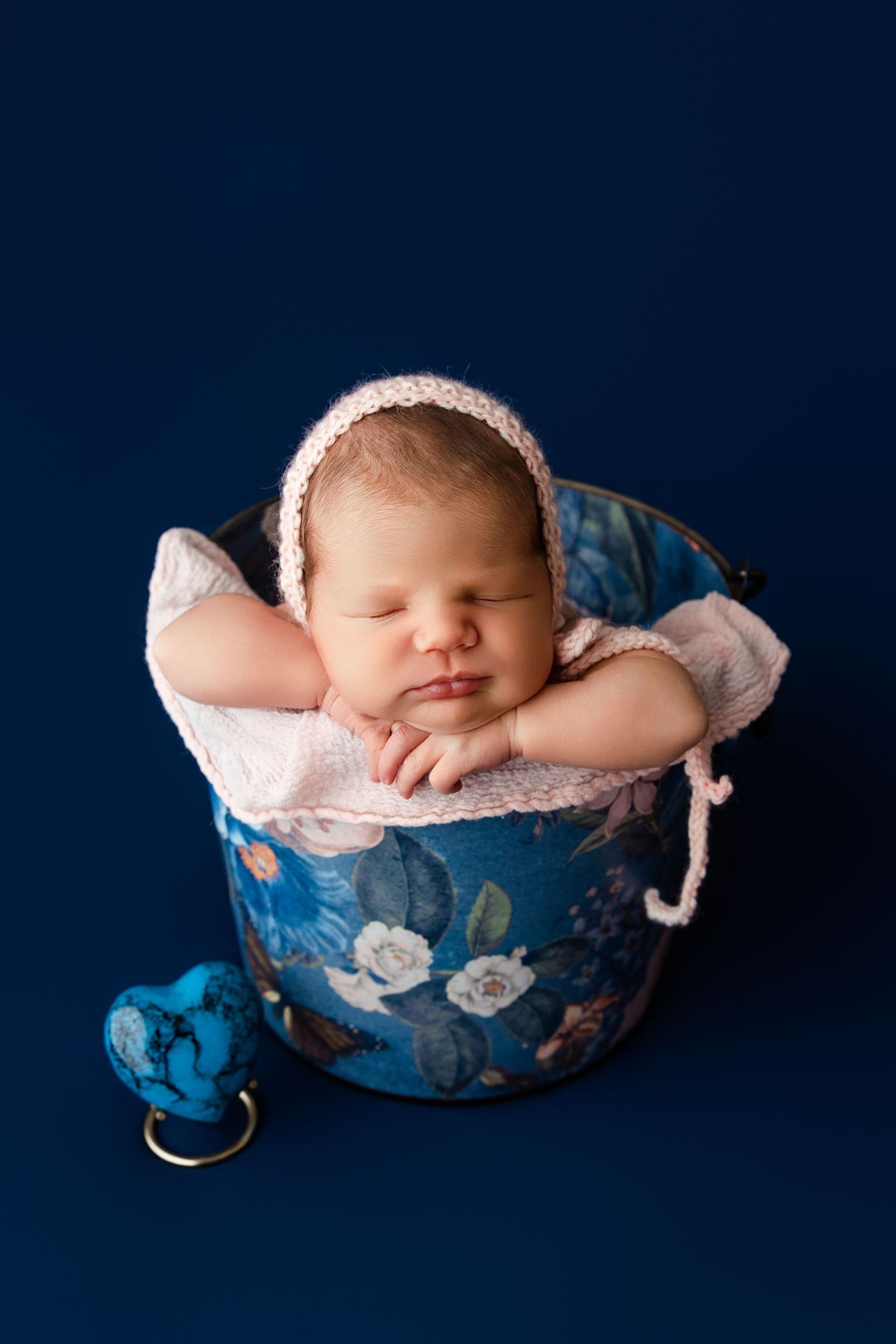 Sleeping newborn in white bonnet rests peacefully in blue floral bucket prop against navy background.
