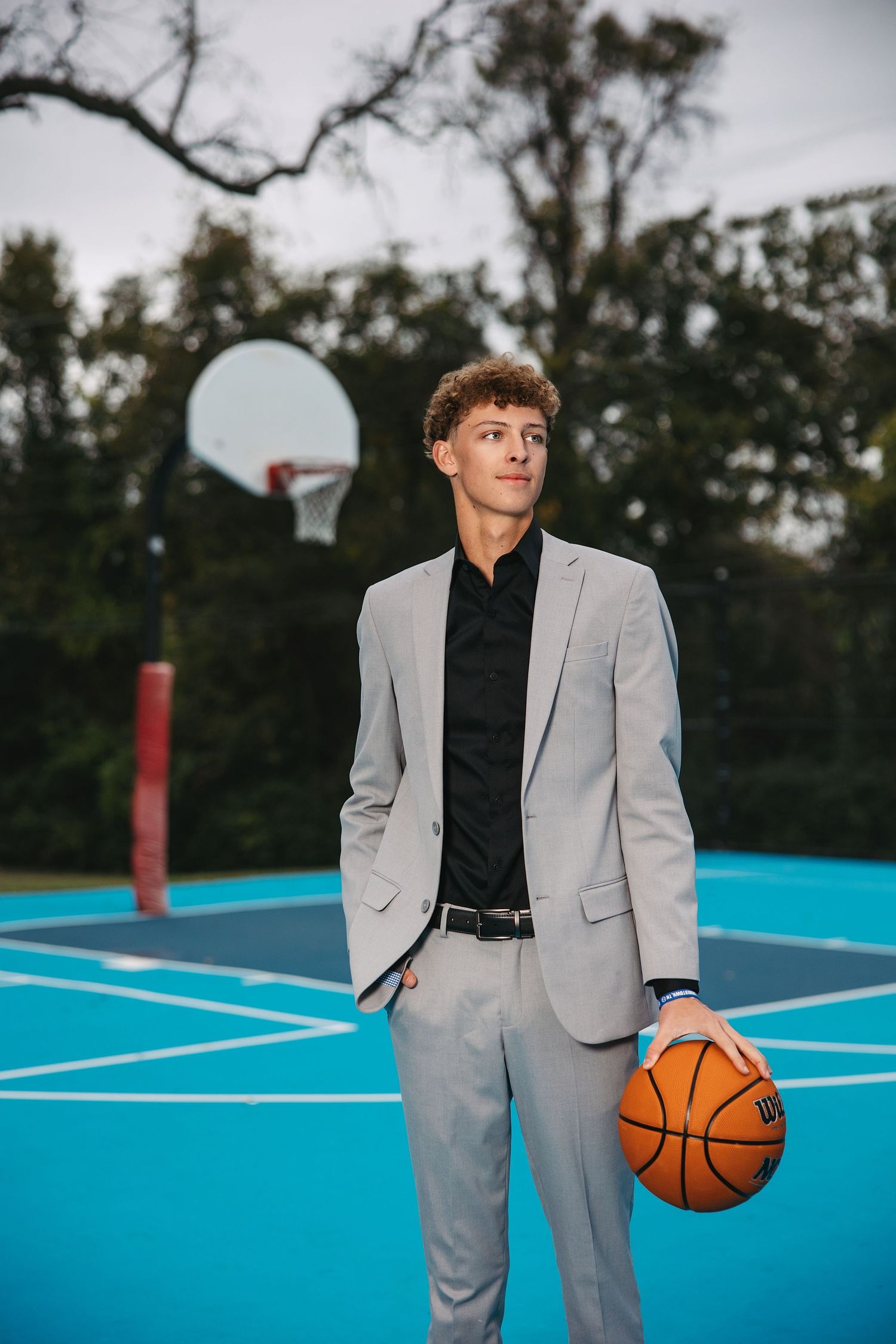 Person in gray suit and black shirt poses with basketball on vibrant blue outdoor court at dusk.