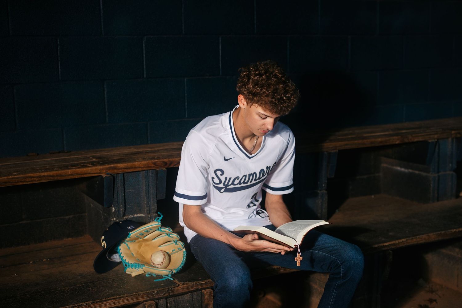 A person in a baseball jersey sits on wooden bleachers reading a book with a baseball glove beside them.