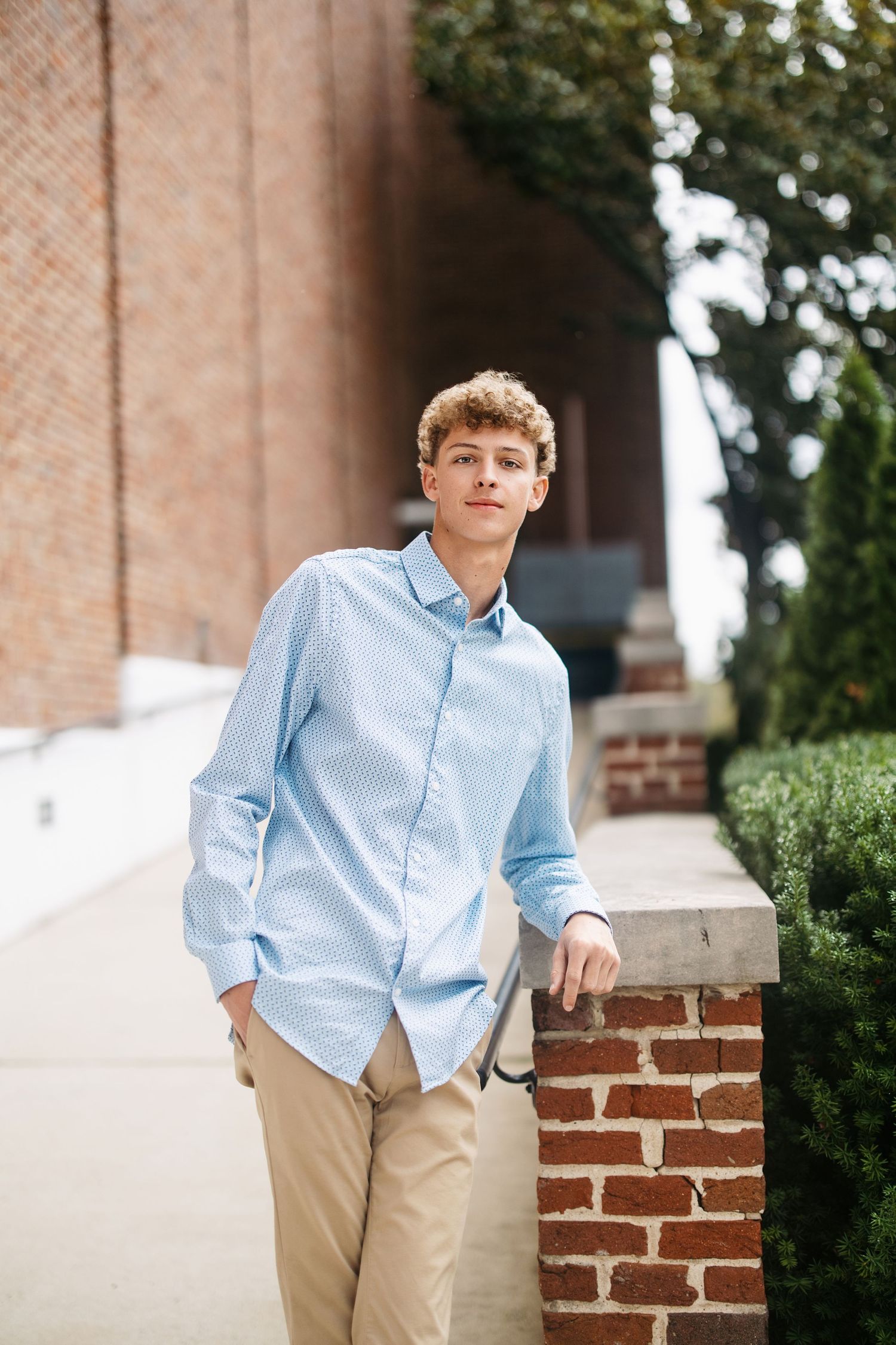A student in a light blue button-down shirt and khaki pants poses casually against a brick wall outdoors.