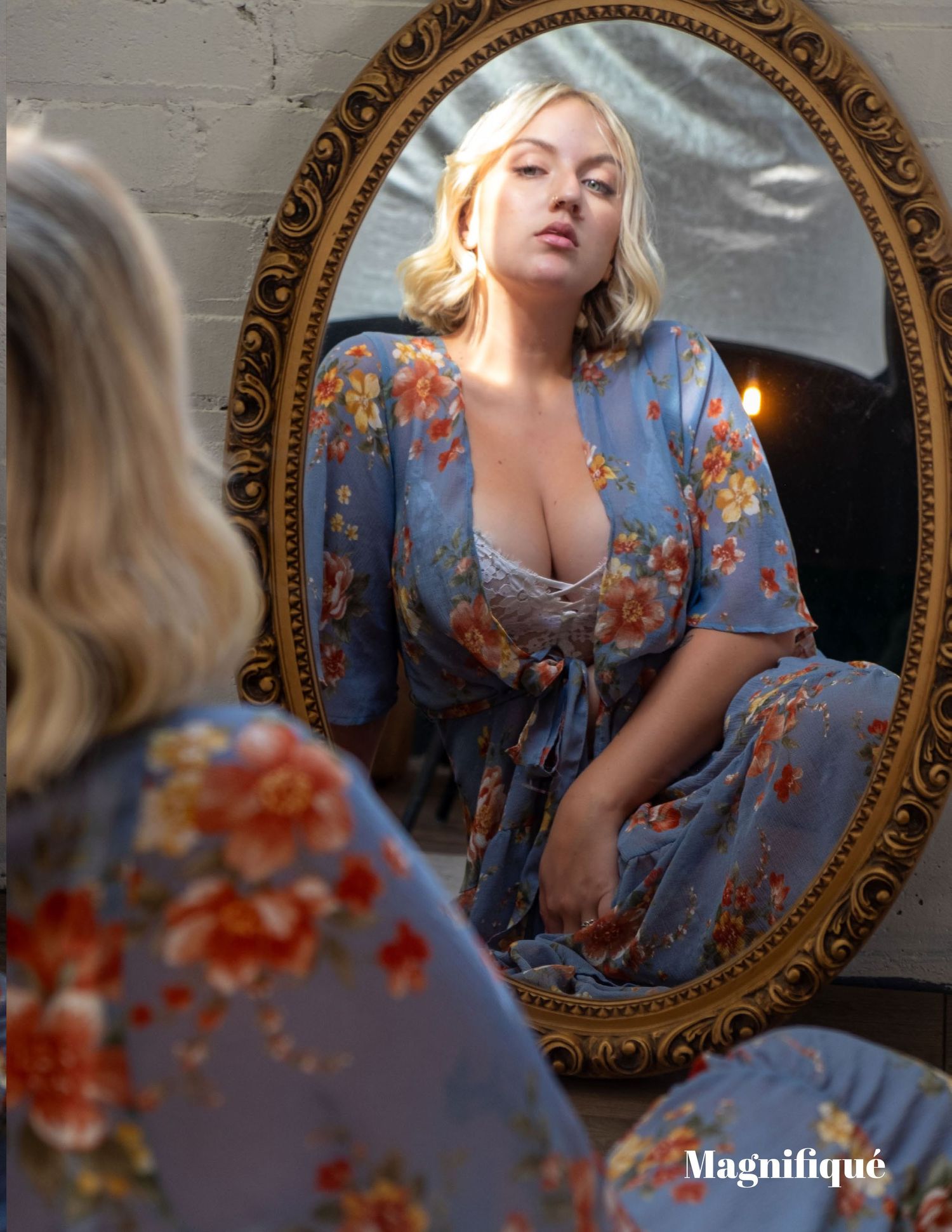 Reflection in ornate gold mirror shows person wearing light blue floral dress with plunging neckline and puff sleeves.