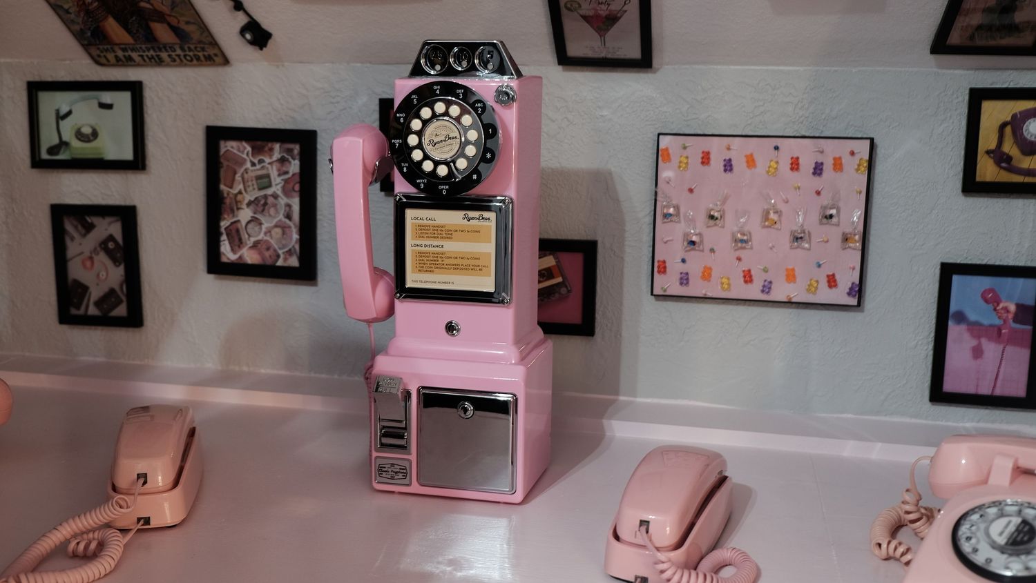 Vintage pink pay phone display with retro pink telephones on white wall with framed art in a museum exhibit.