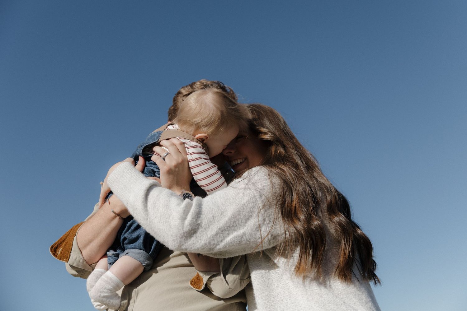 A mother tenderly embraces her young child under a clear blue sky outdoors.