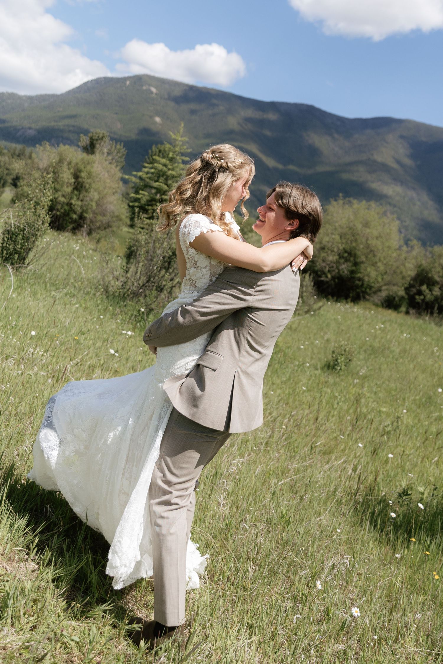 Groom lifts bride in white dress on green mountain meadow, sharing a romantic moment on their wedding day.