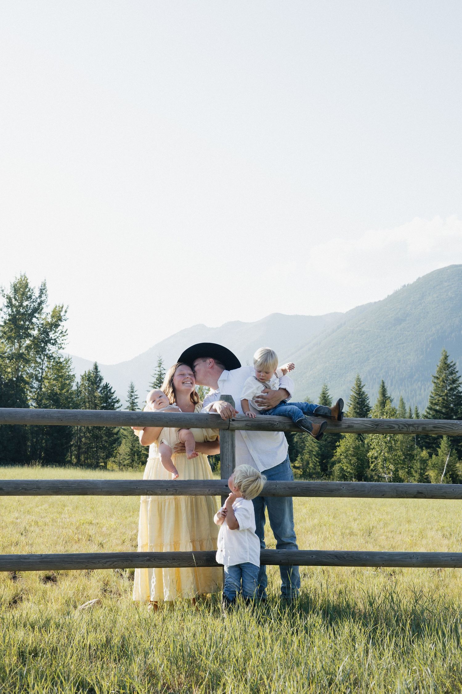 Family enjoys a tender moment together by a wooden fence in a scenic mountain meadow at sunset.
