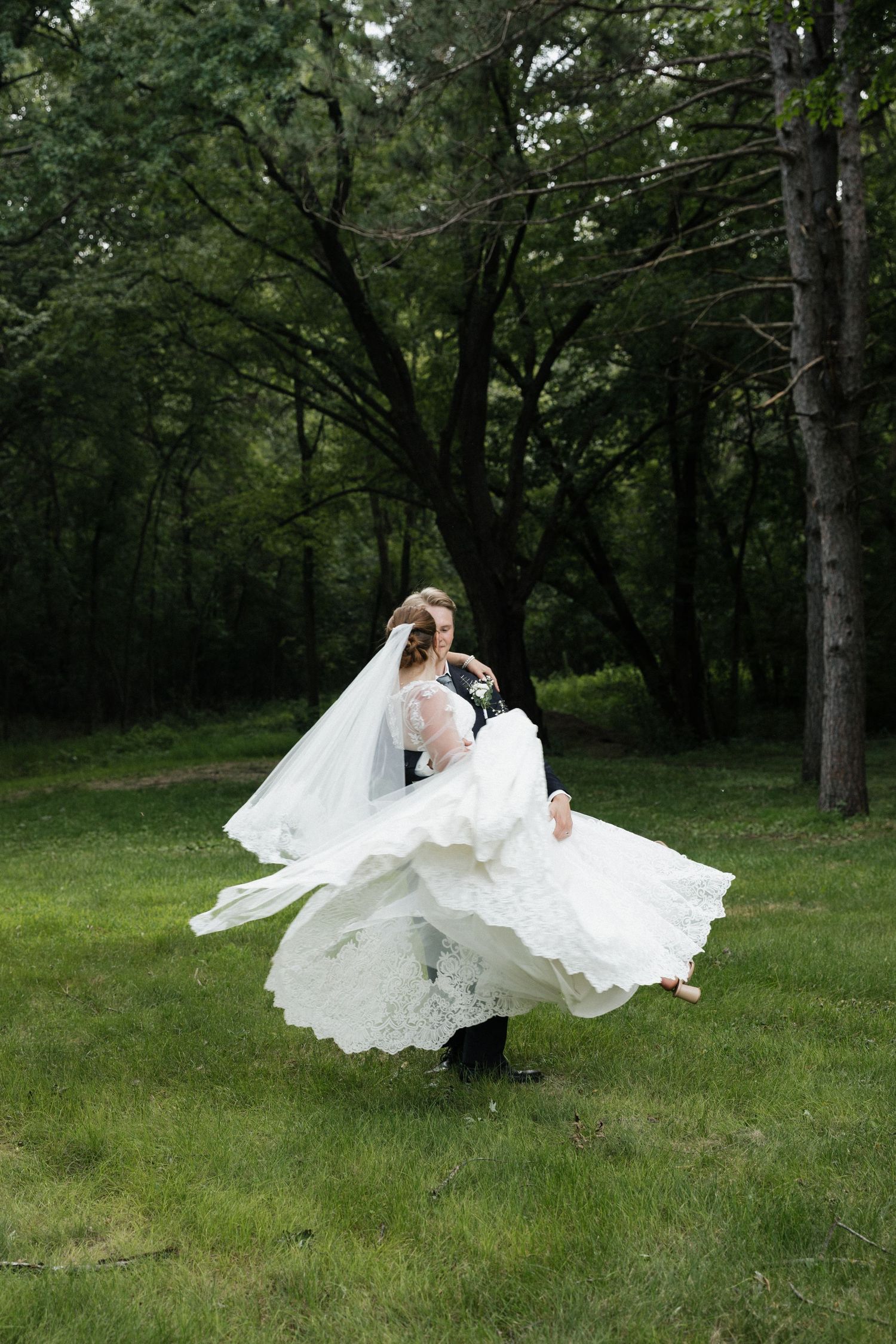 A couple in wedding attire twirls gracefully across a lush green lawn surrounded by trees.