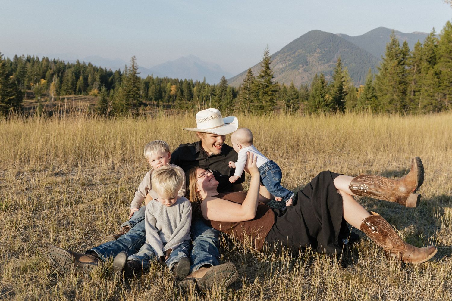 A romantic couple lounges together in a grassy meadow with mountains and evergreen trees in the background.