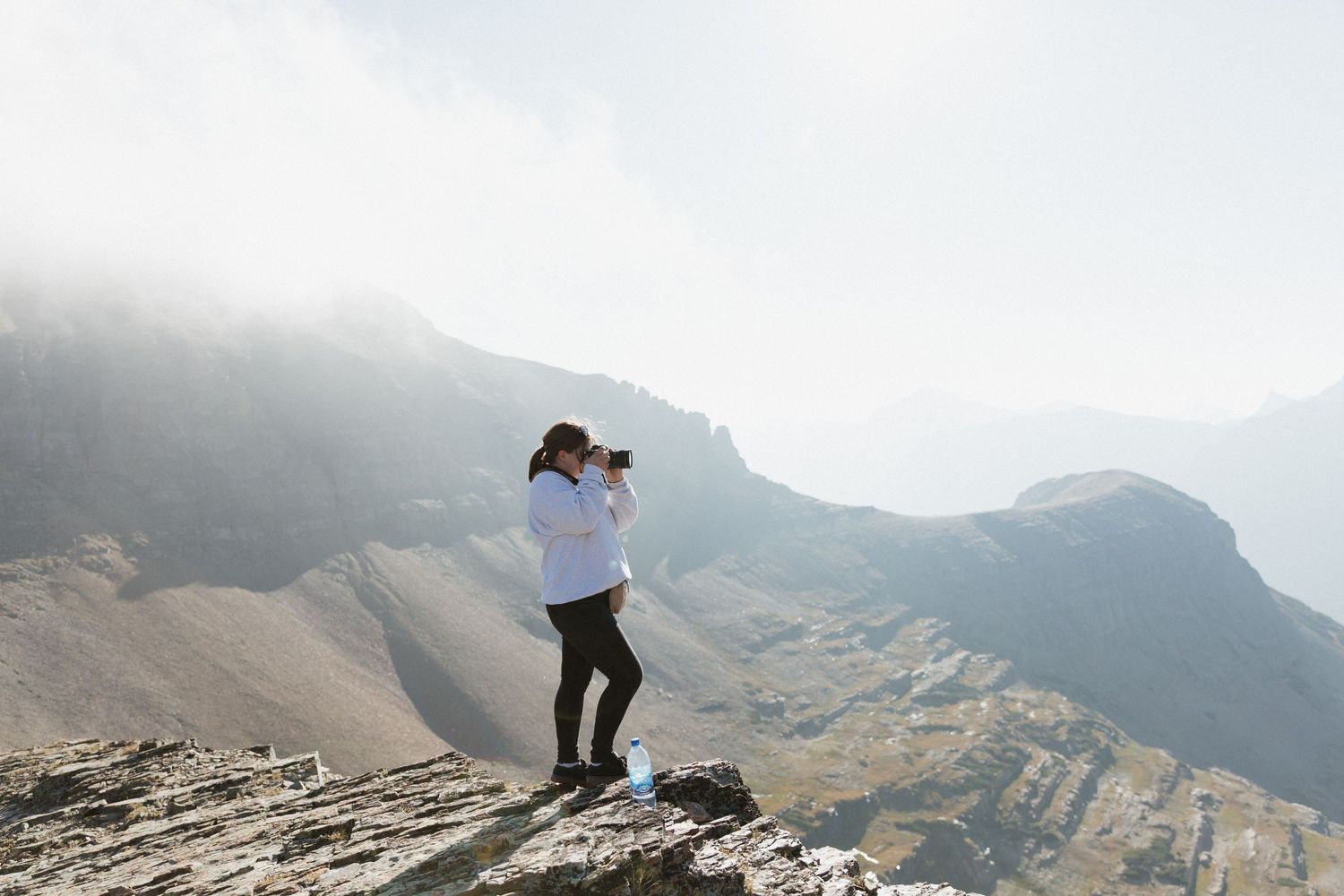 Photographer in white sweater captures mountain landscape from rocky summit on misty morning.