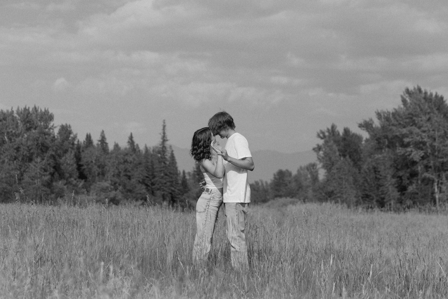 A black and white photograph of a romantic couple embracing in a grassy meadow with trees on the horizon.