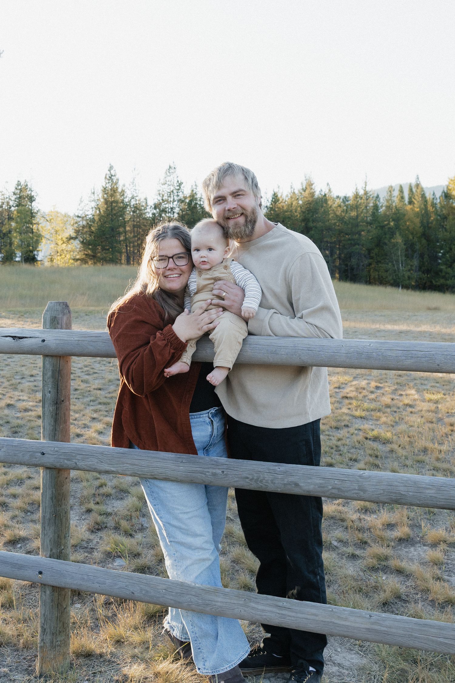 Family shares tender moments together while leaning against a rustic wooden fence in a rural field at sunset.