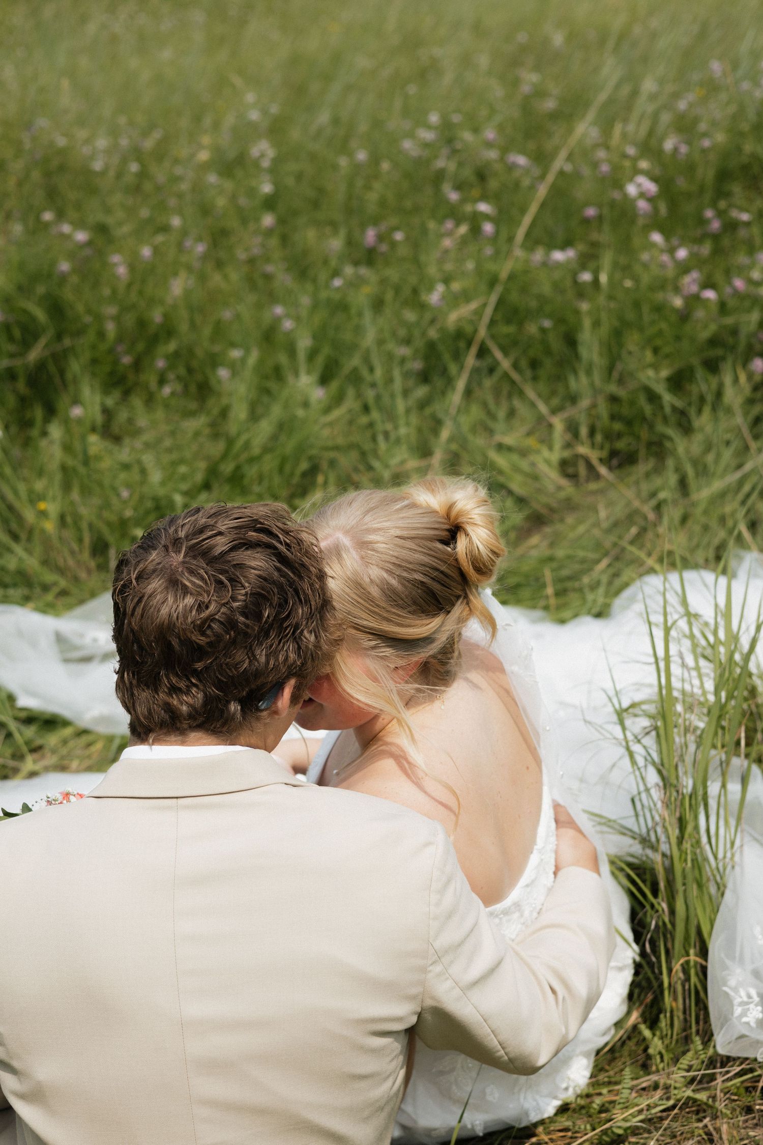 A couple in wedding attire sits together in a wildflower meadow on a sunny summer day.