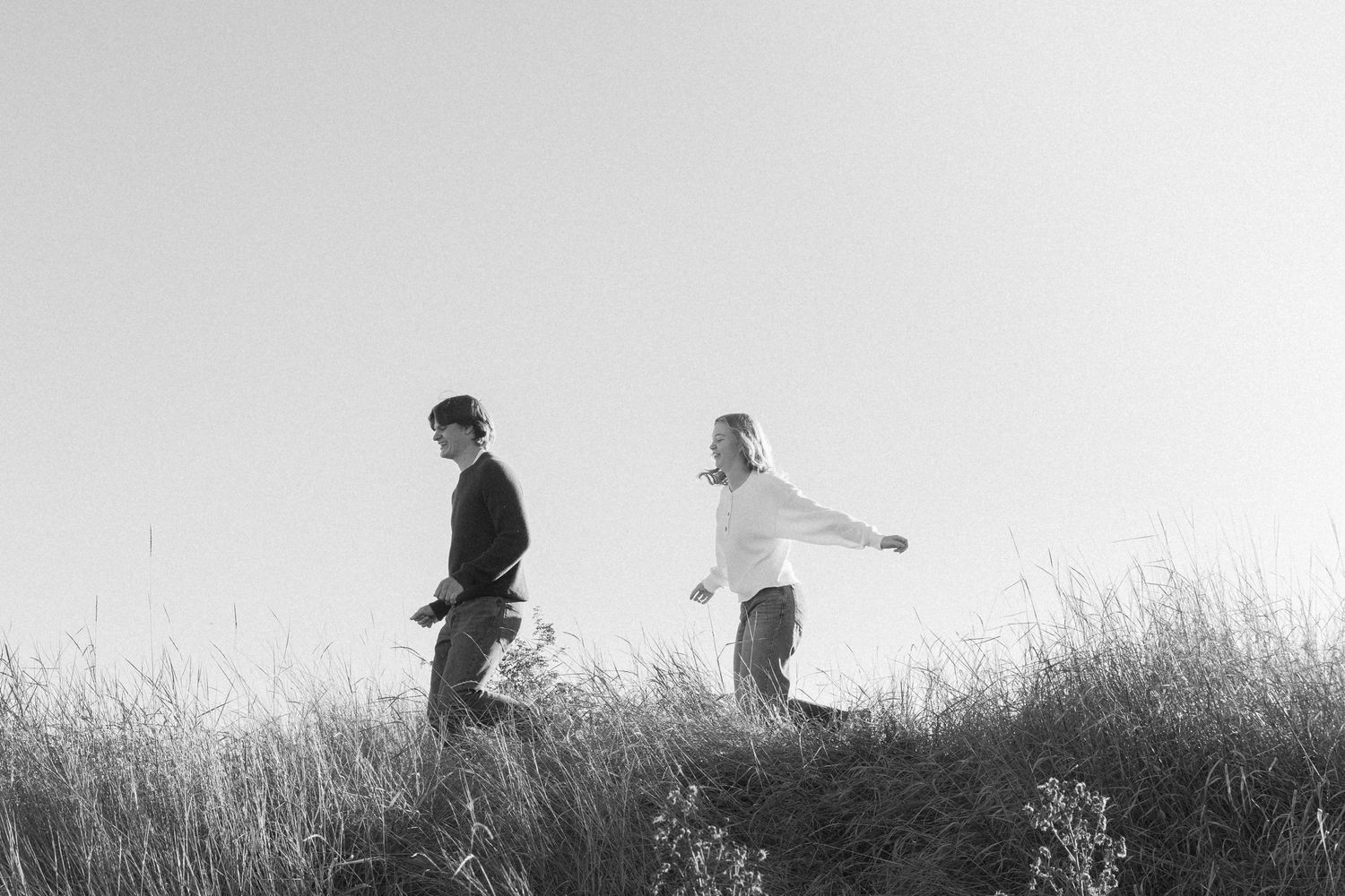 Two silhouetted figures walk through tall grass against a bright sky background in a black and white photograph.