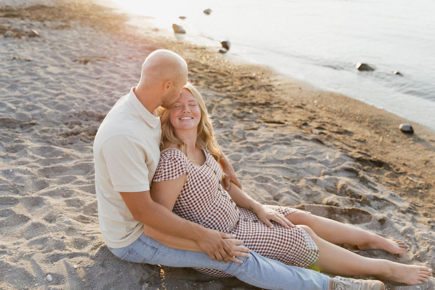 Happy couple sitting on a sandy beach at sunset, woman leaning against man, both smiling warmly.