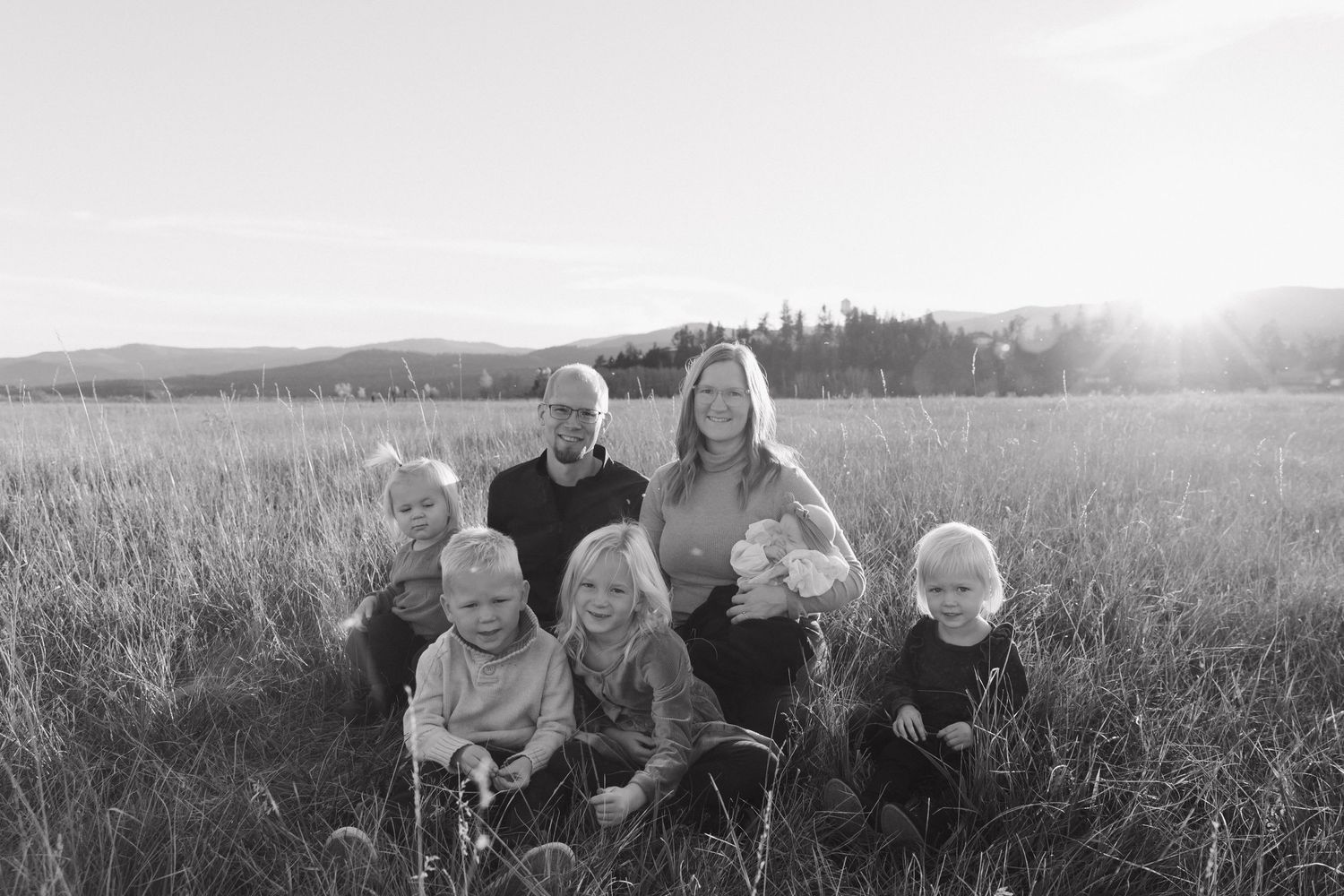 A family sits together in a sunlit field of tall grass during golden hour, captured in black and white.