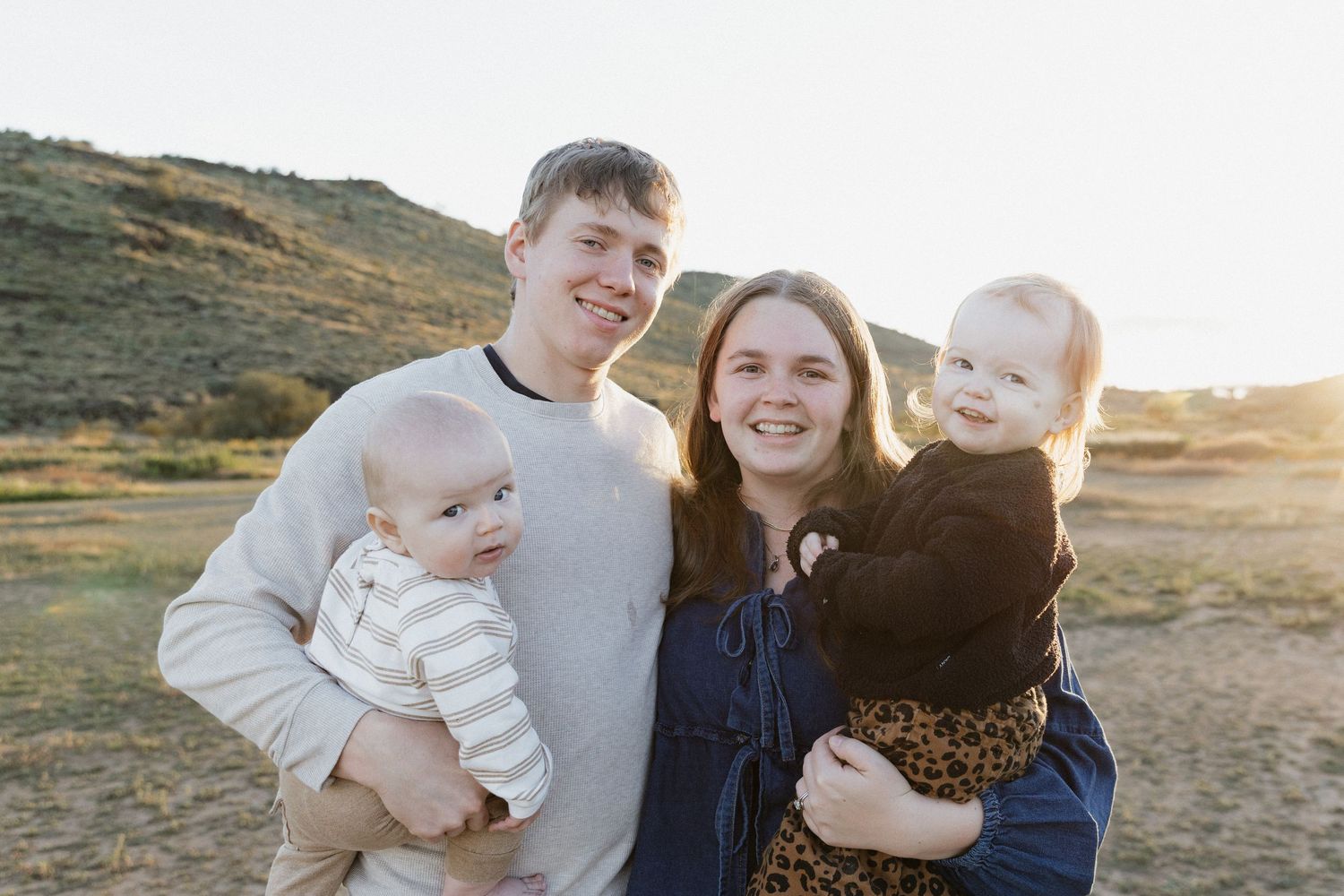 A family of four poses together outdoors during golden hour with hills in the background.