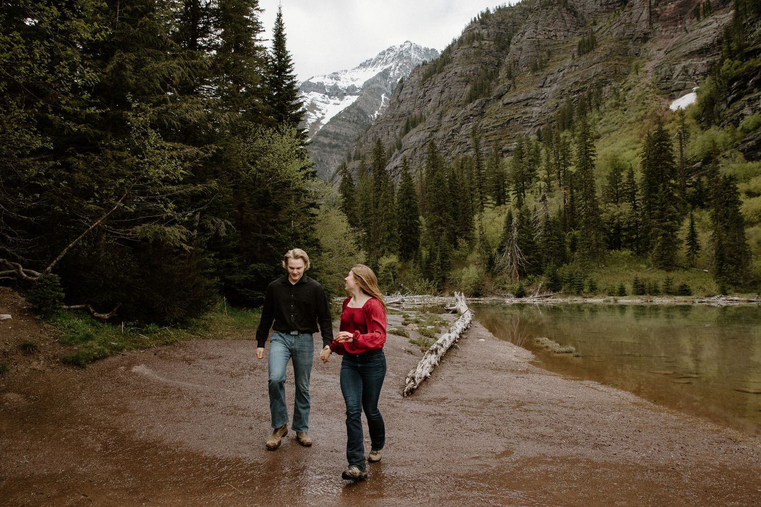 Two people walking on a scenic mountain trail alongside a serene lake with evergreen trees and snow-capped peaks.