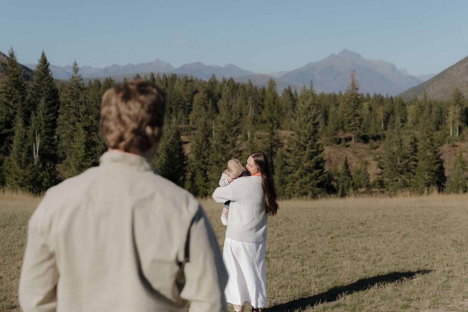 Couple embracing in a mountain meadow surrounded by pine trees, with a photographer in the foreground capturing the moment.