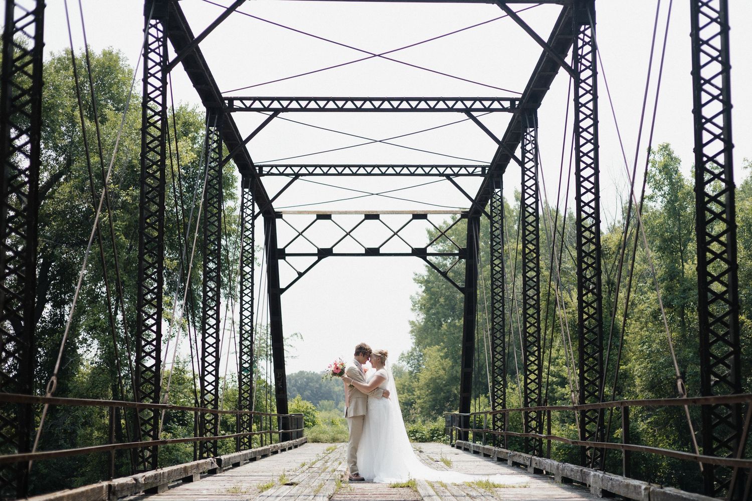 Two people walk hand in hand on a historic steel truss bridge surrounded by lush green trees and natural scenery.