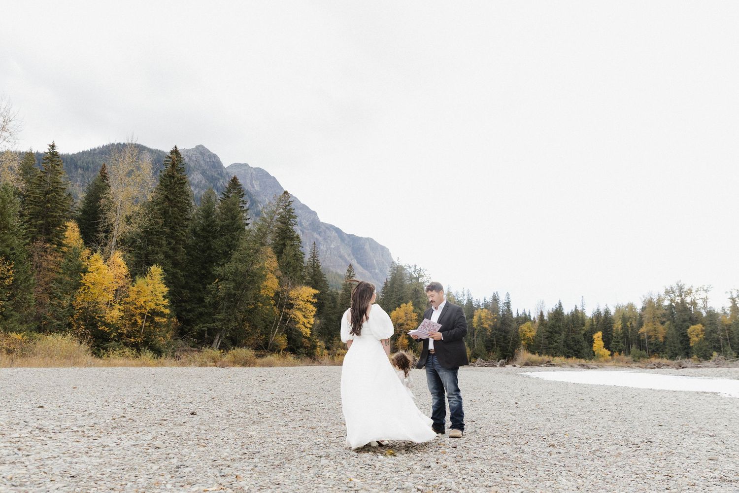 A couple in wedding attire stands together against a backdrop of mountains and autumn trees in a scenic outdoor setting.