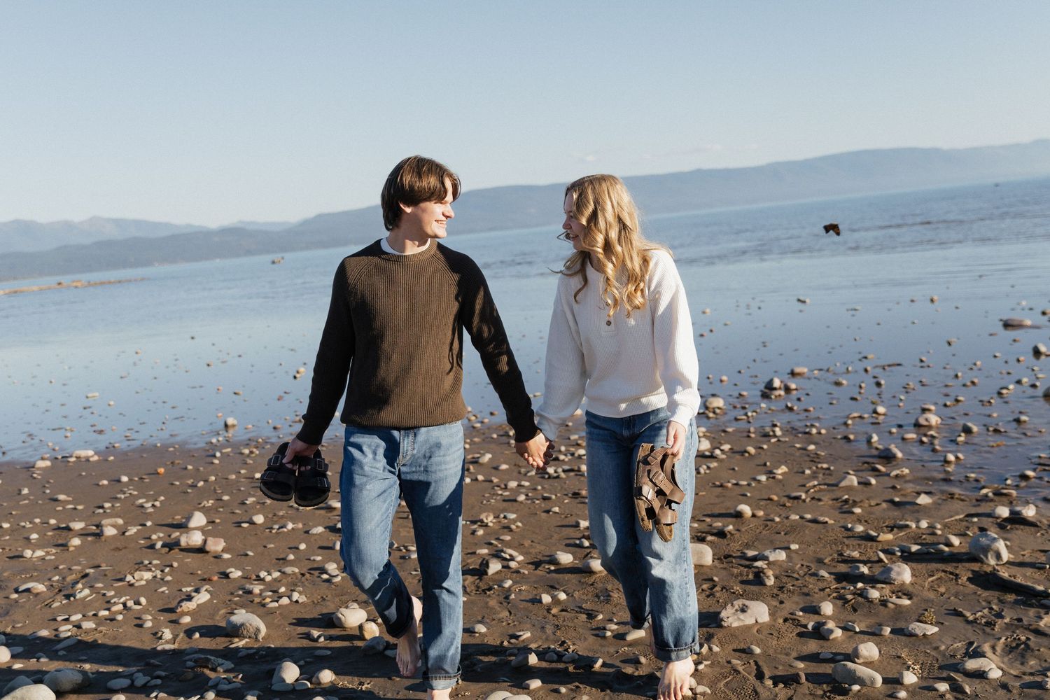 Two people walk hand in hand along a rocky beach with mountains and water in the background on a sunny day.