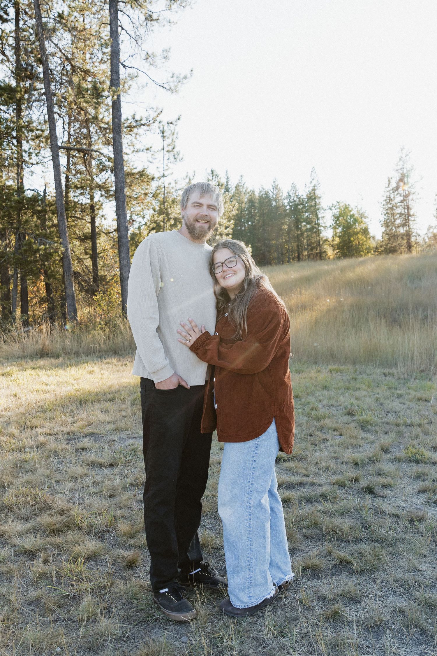 A couple embraces in a sunlit meadow surrounded by tall pine trees on an autumn afternoon.