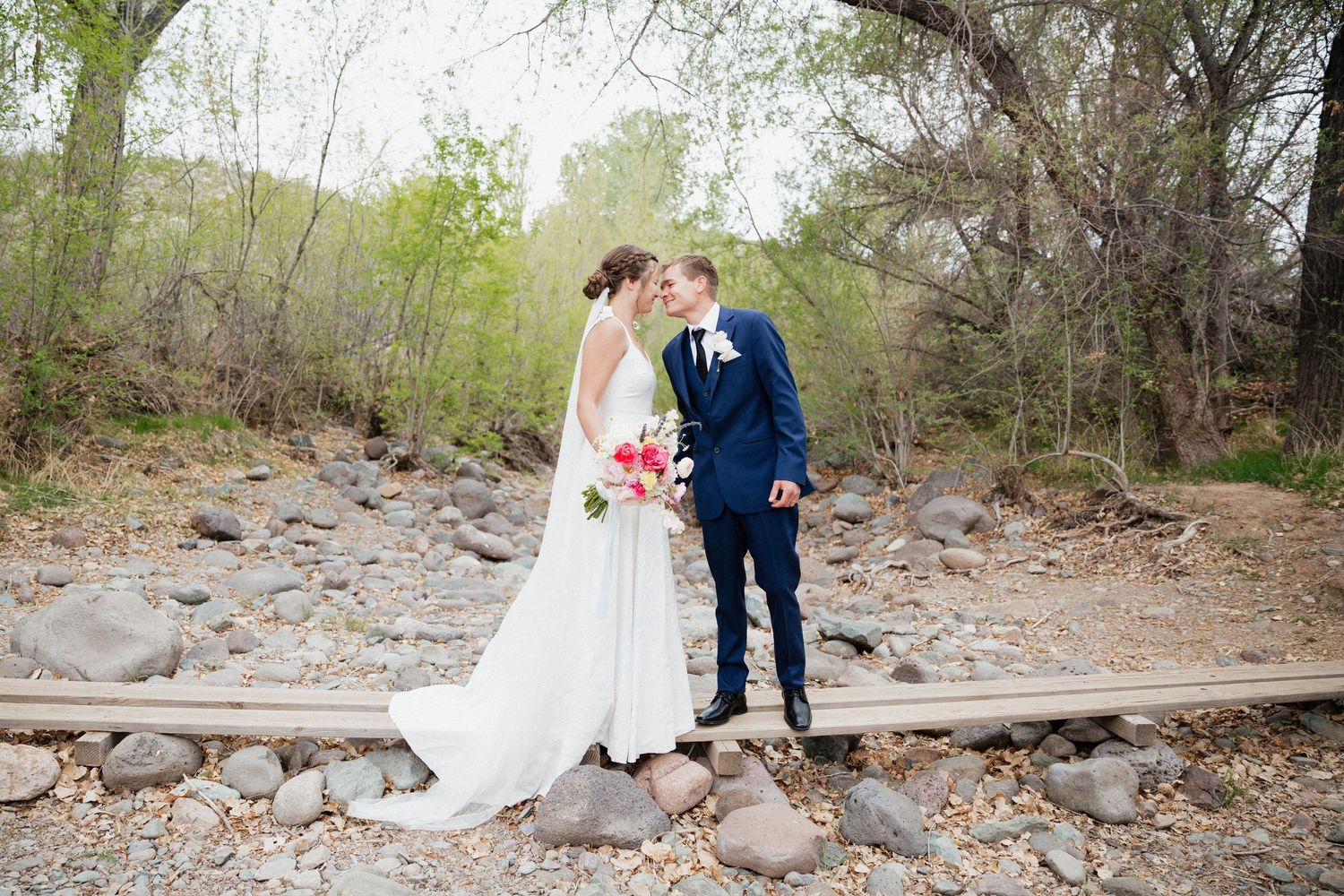 Two people in wedding attire stand together on a rocky riverbank beneath willow trees in a natural outdoor setting.
