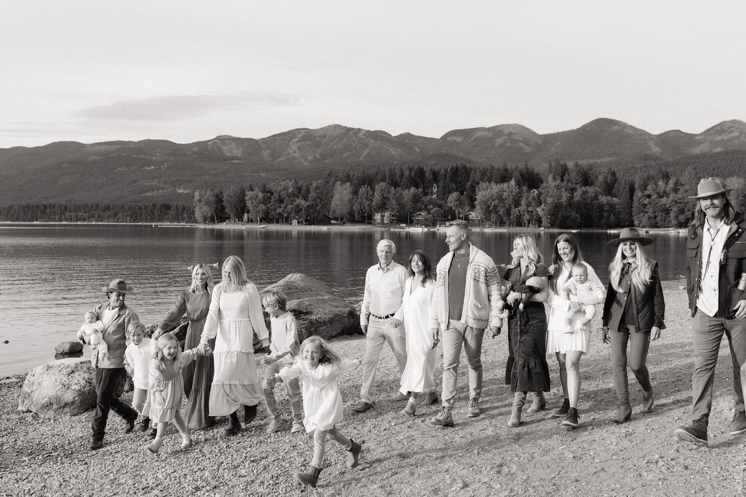 Black and white photo of a large family group walking along a lakeside beach with mountains in the background.
