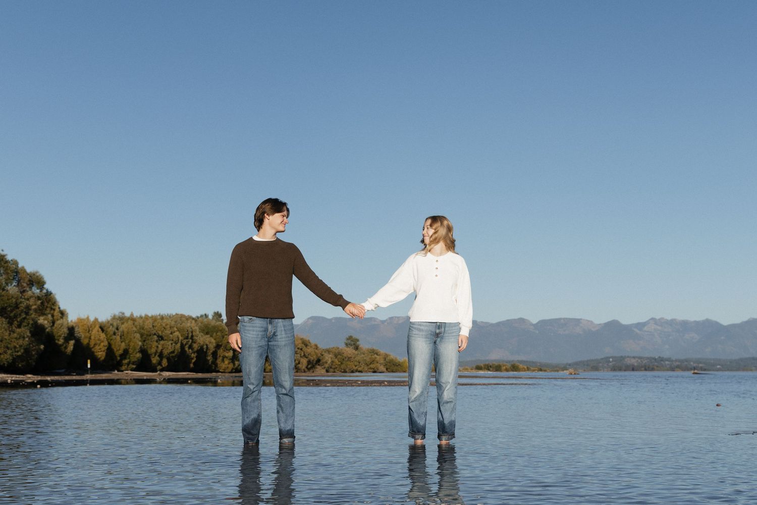 Couple holding hands while standing in shallow lake water with mountains and clear blue sky backdrop.