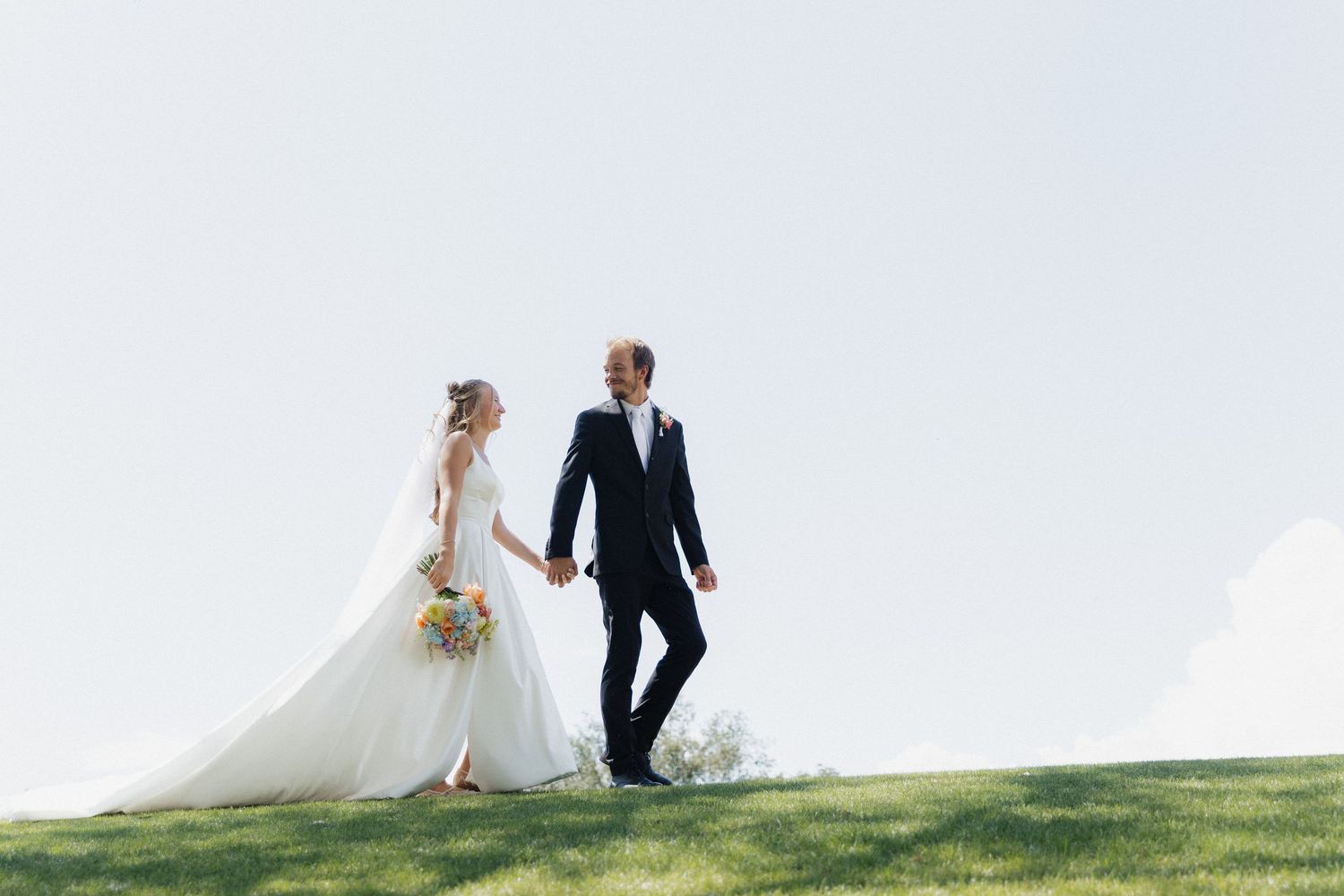 Bride and groom holding hands walking on green grass hill in wedding attire, bride carrying colorful bouquet.