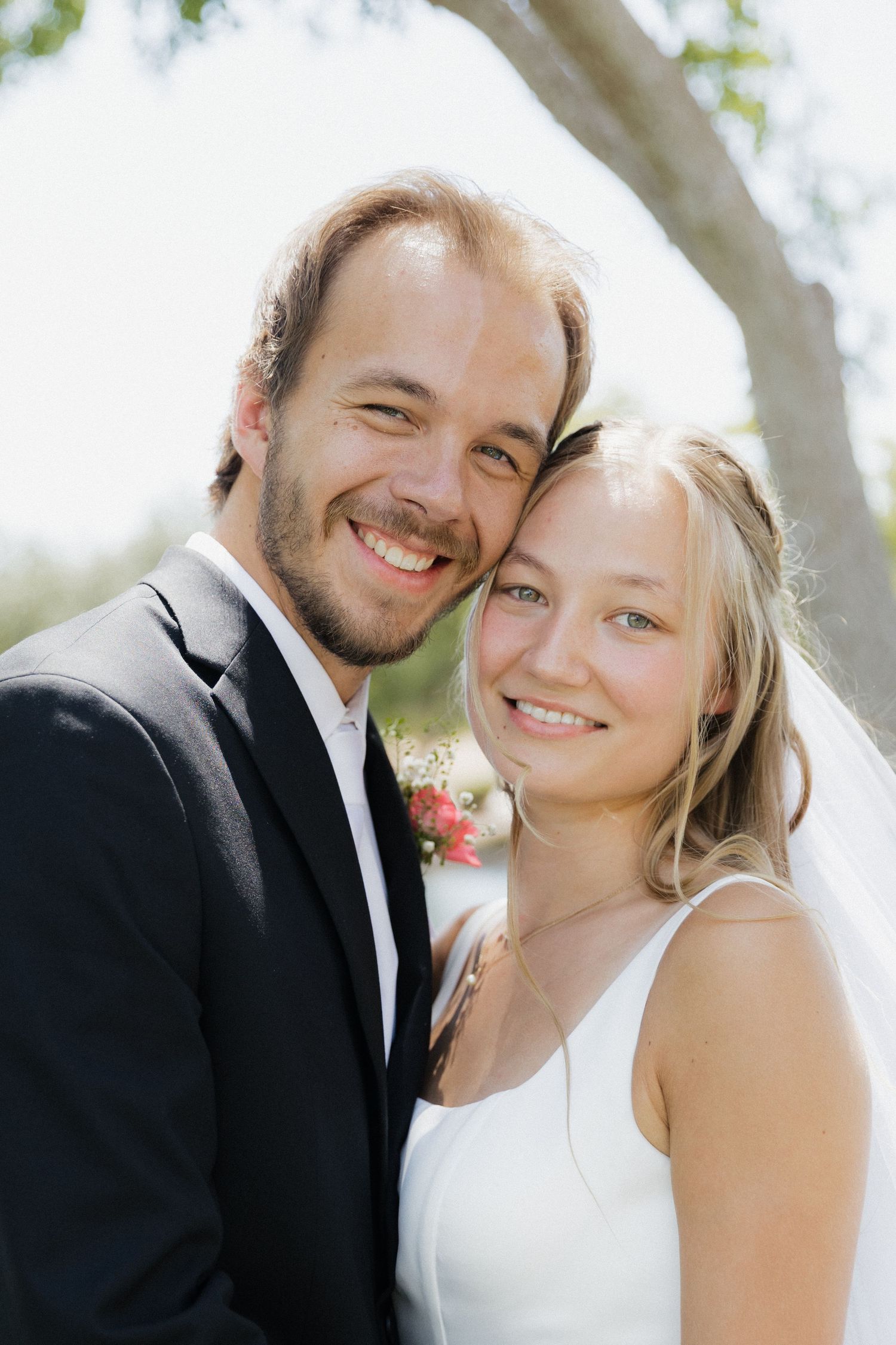 Smiling bride and groom pose cheek to cheek outdoors on their wedding day, wearing white dress and black suit.