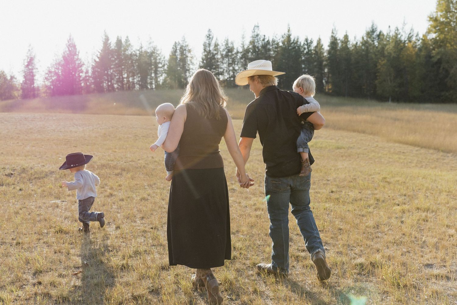 A family walks together in a sunlit meadow surrounded by evergreen trees on a warm summer evening.