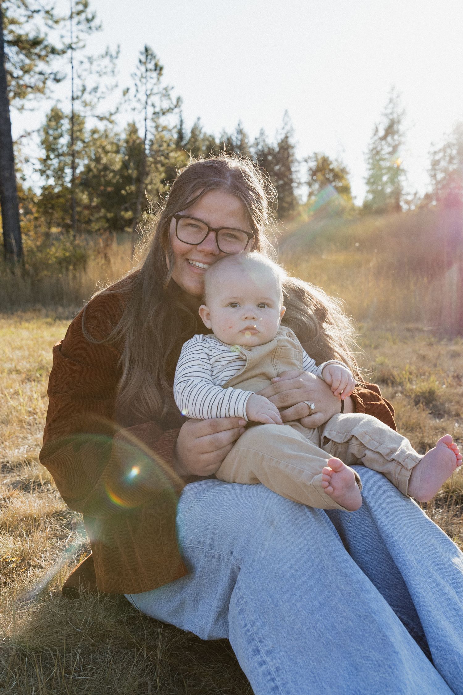 Parent and baby share tender moment while sitting together on blanket in sunlit outdoor setting.
