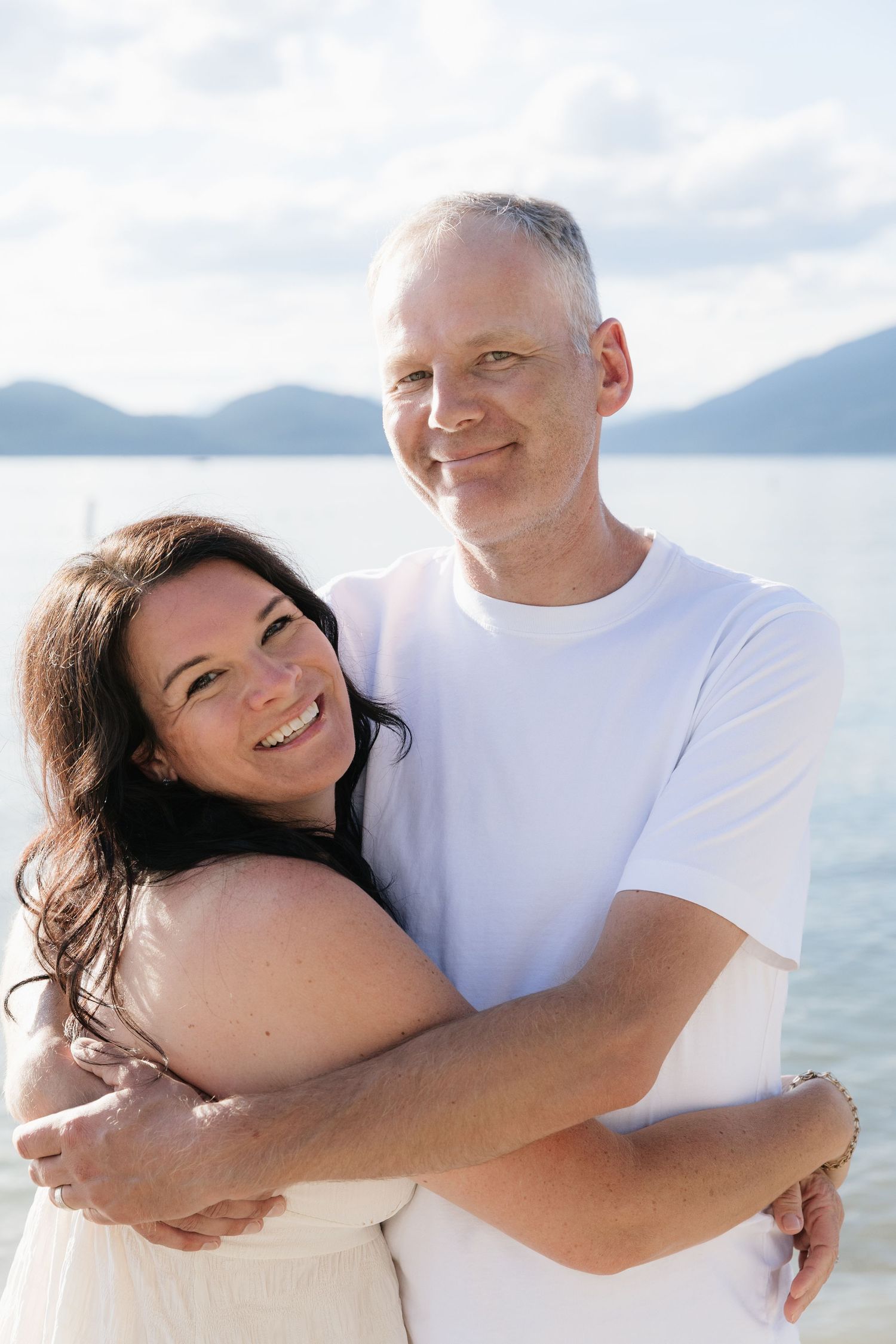 Happy couple embracing on a waterfront with mountains in the background on a sunny day.