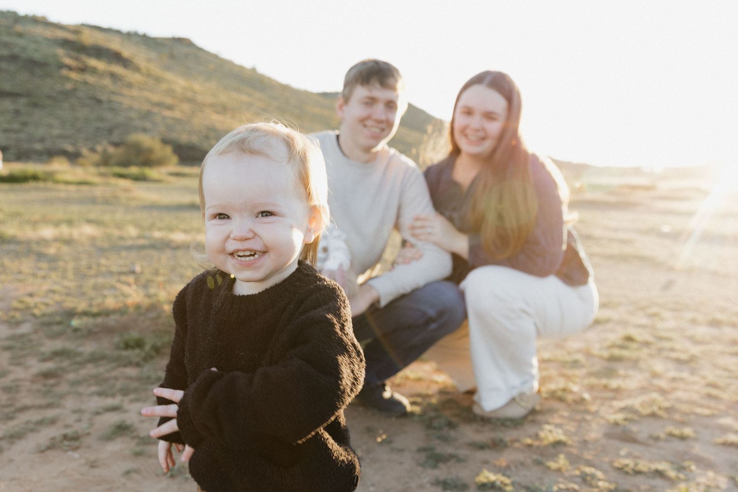 A happy toddler smiles outdoors while young parents crouch behind on sandy terrain with golden hills in the background.