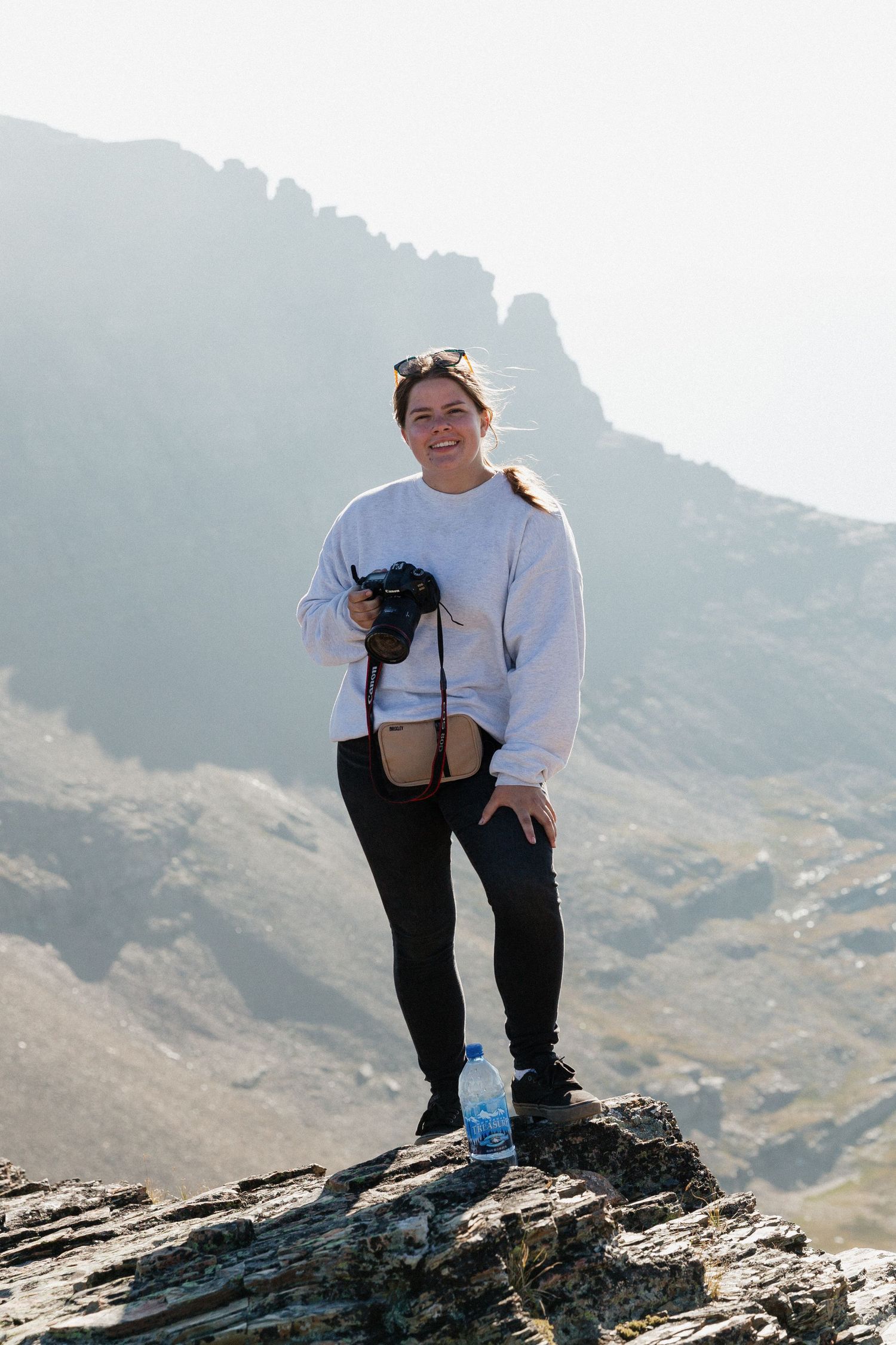 A hiker in a white sweatshirt stands on a rocky mountain summit with their small black dog against a misty backdrop.