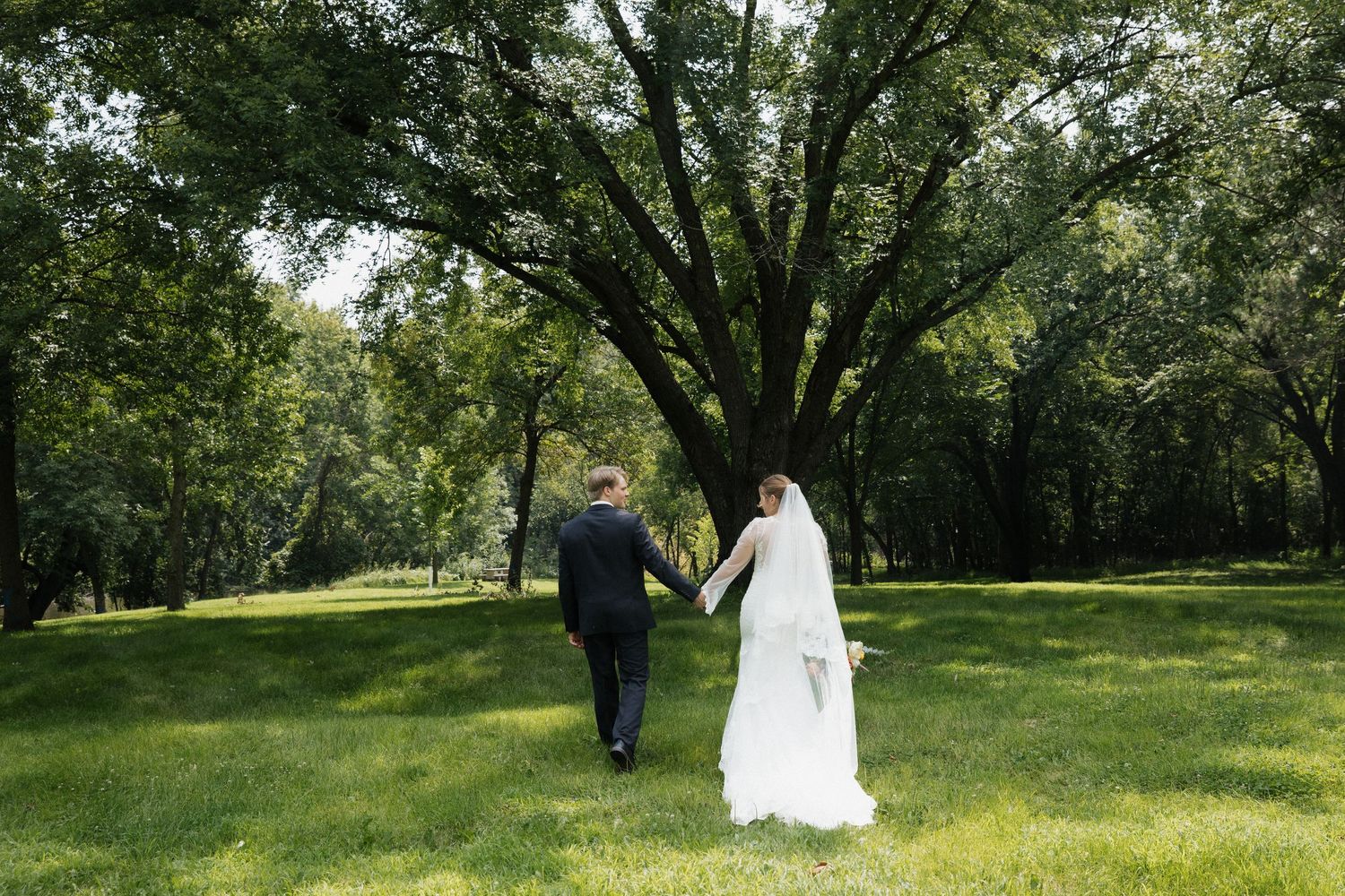 A person in a white wedding dress walks through a lush green park setting during a photo session.