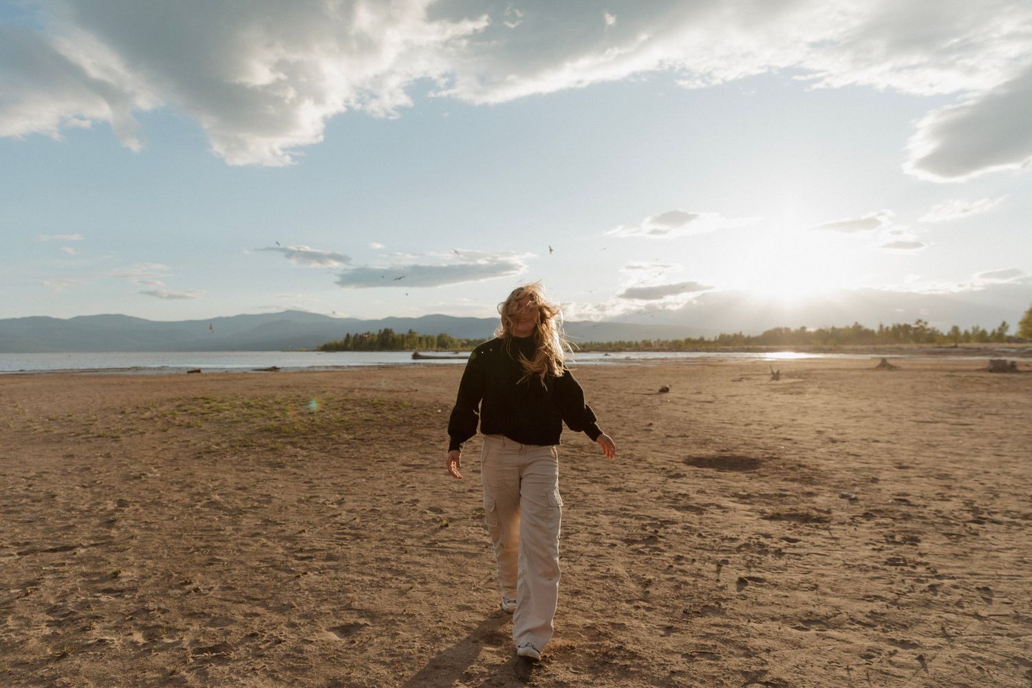Someone spins joyfully on an empty beach at sunset with mountains and dramatic clouds in the background.