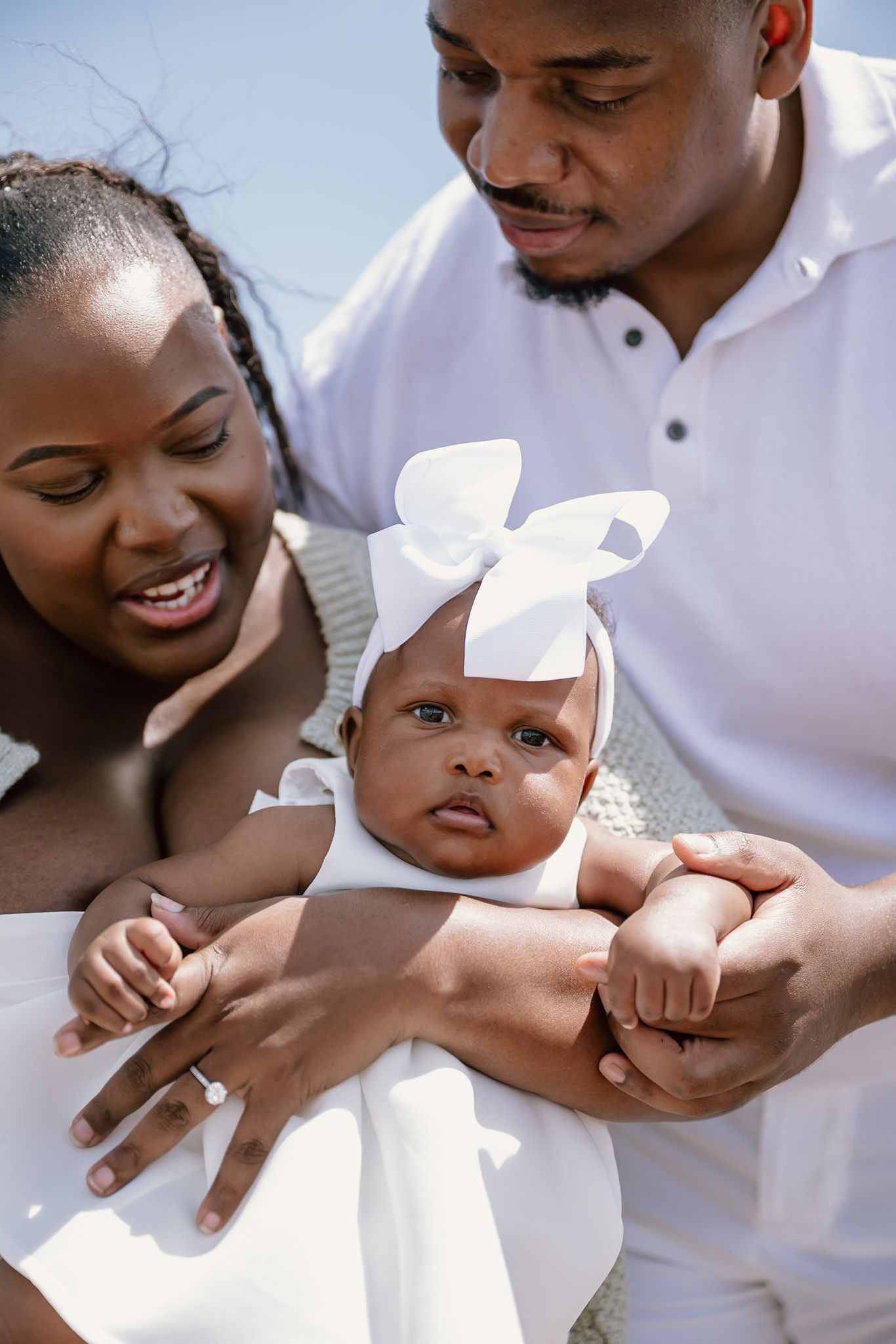 Happy Black family with parents holding their newborn baby girl wearing a white dress and bow headband outdoors.