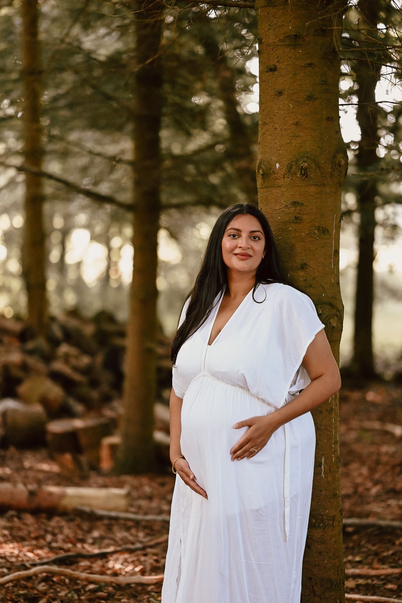 A person in a flowing white dress poses for maternity photos in a serene forest setting with golden sunlight.