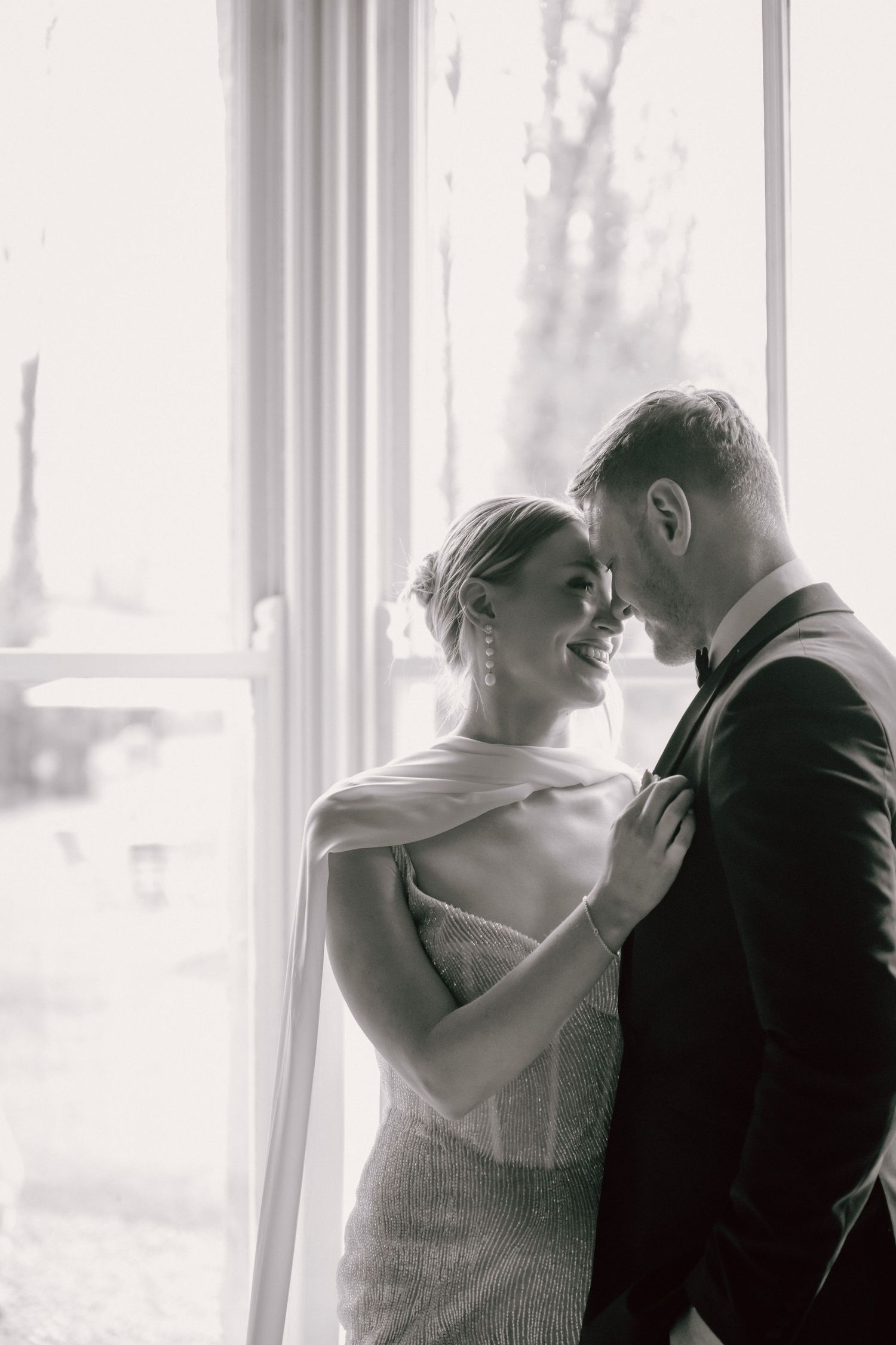 A romantic black and white wedding photo of a couple embracing by a bright window in an intimate moment.