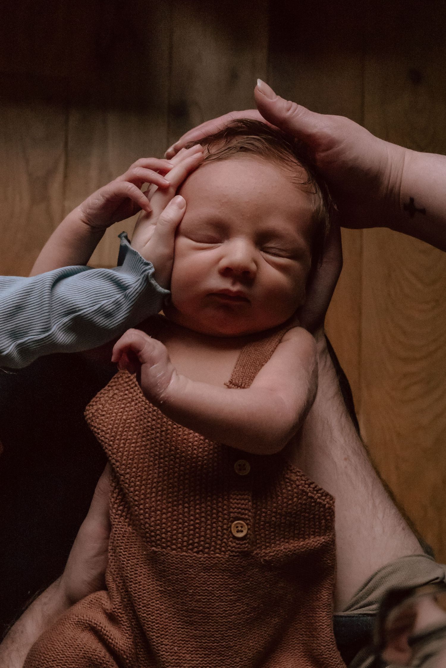 Newborn baby in brown knit sweater sleeps peacefully while gentle hands cradle their head.