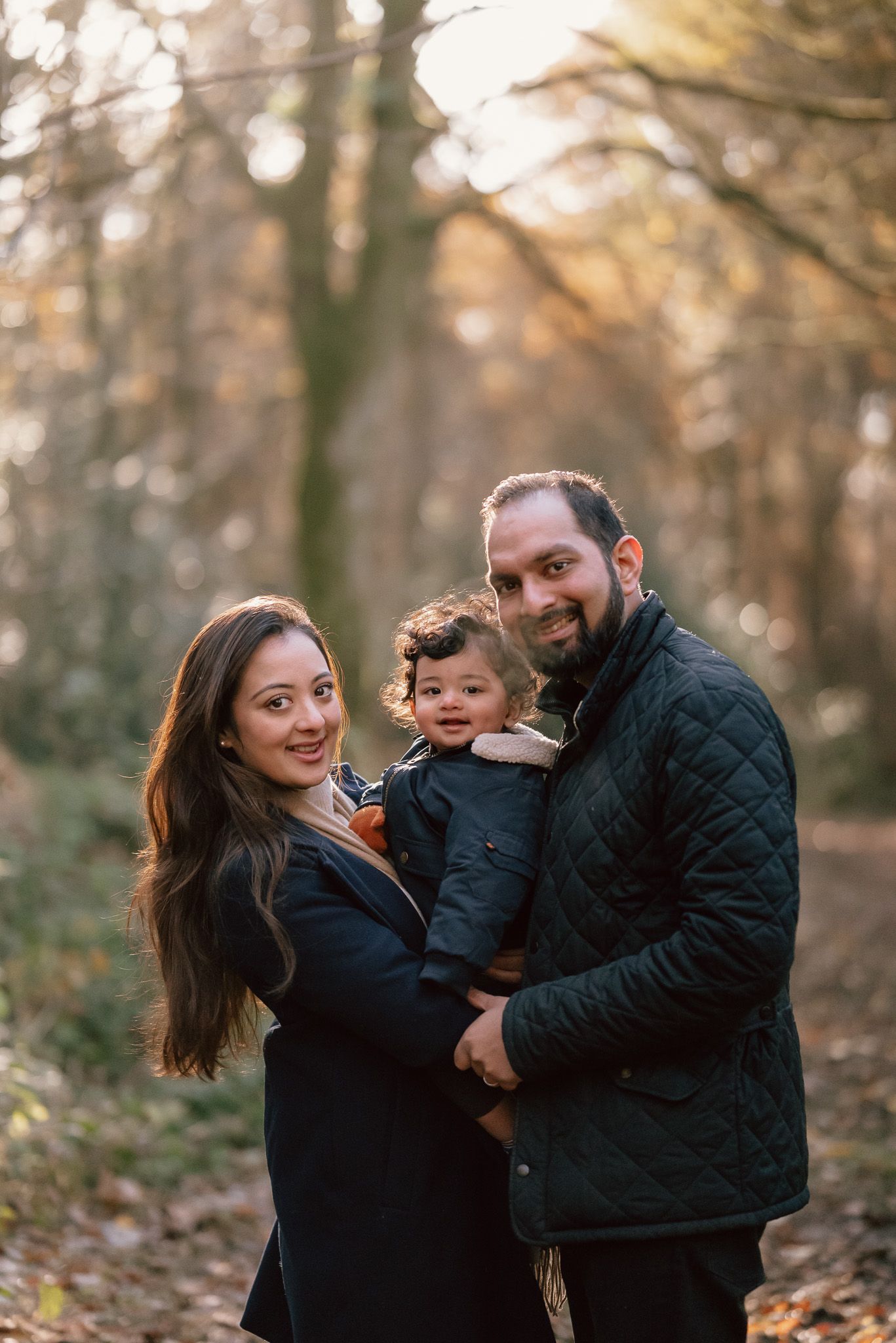 A family poses together on an autumn day in a wooded area with warm golden light filtering through the trees.